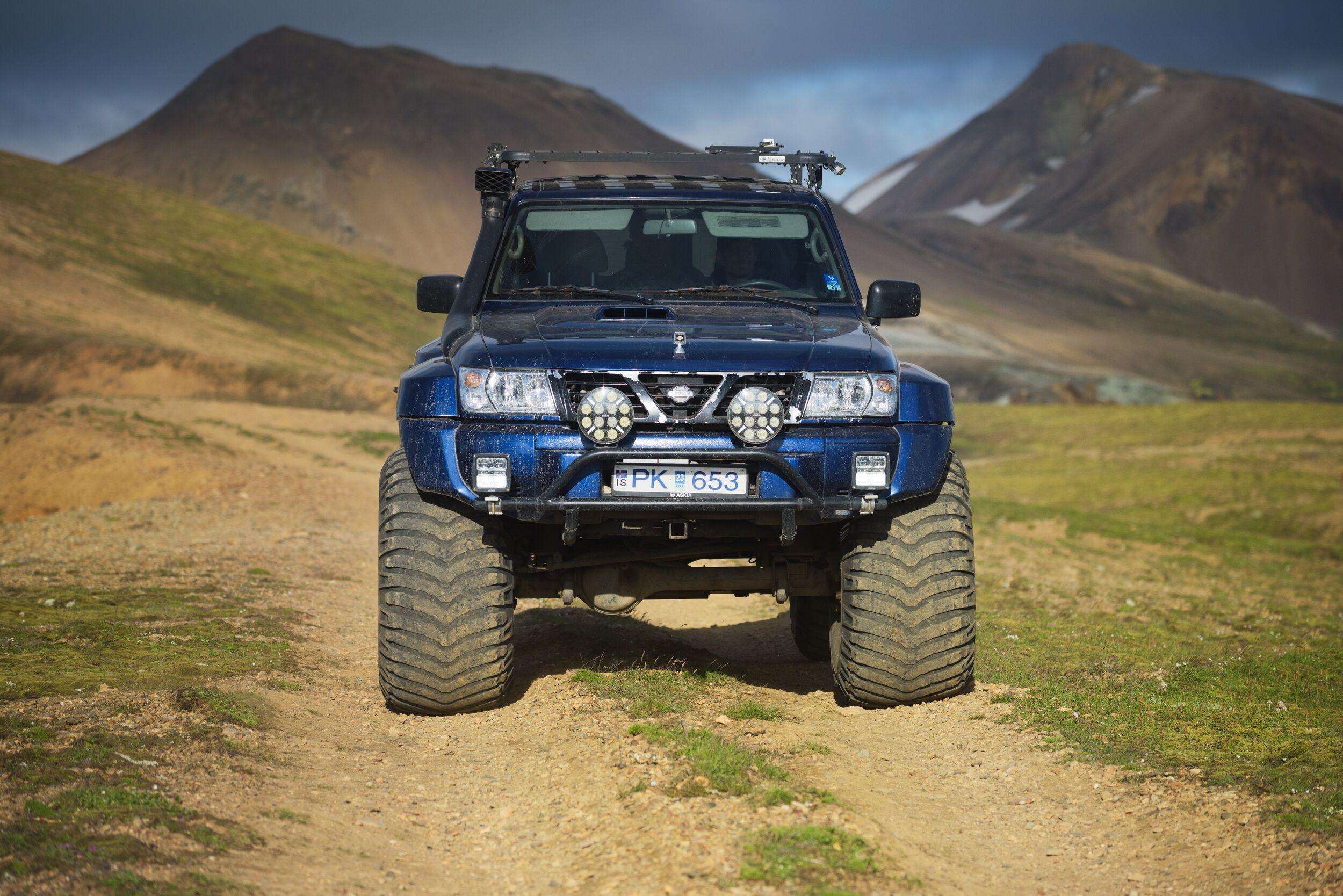 A dark blue modified Nissan Patrol 4x4 with huge off-road tires and auxiliary lights, parked on a dirt track in a rugged mountainous landscape.