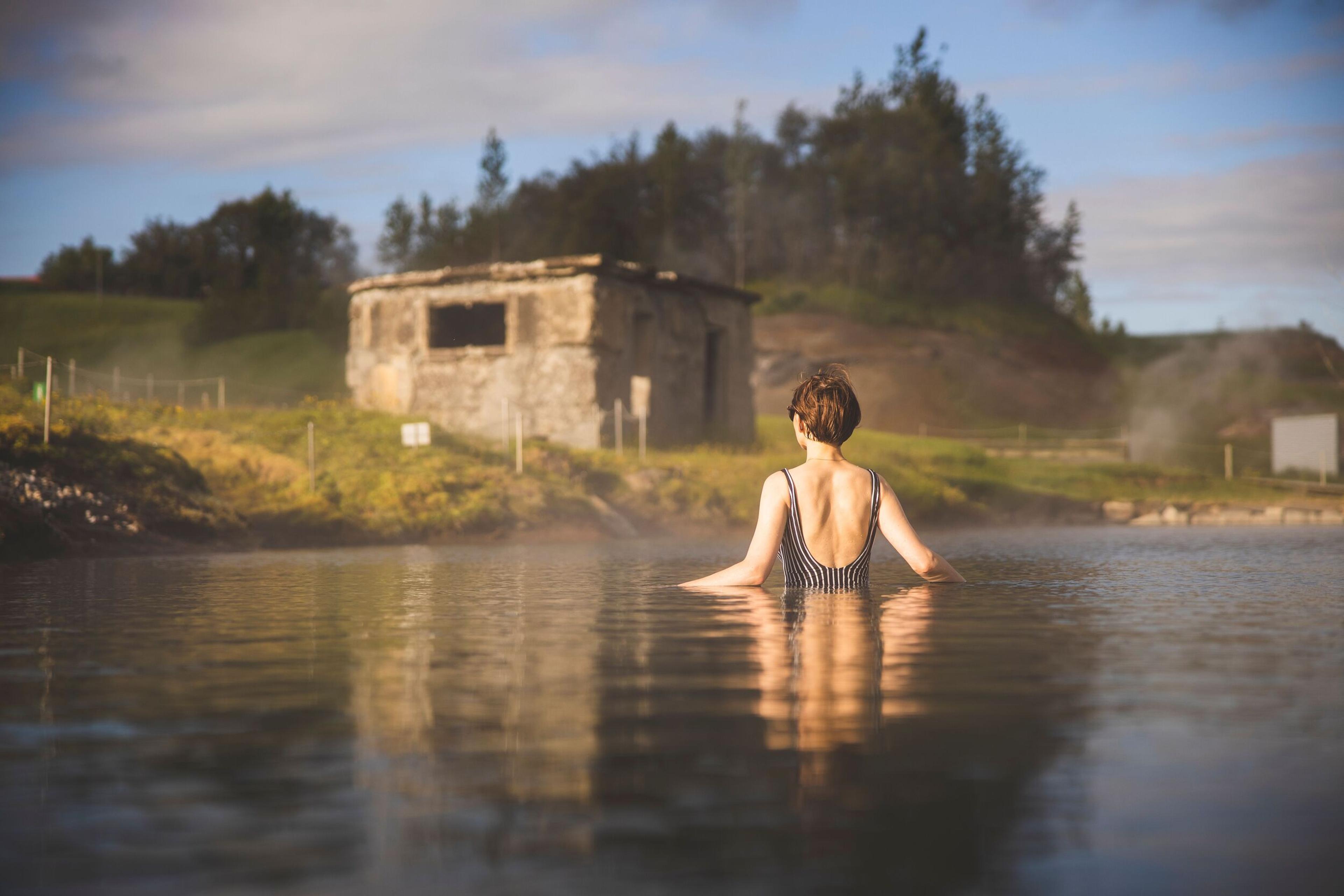 A person in a striped swimsuit soaks in a steaming hot spring, facing away from the camera, with a rustic building on the shore.