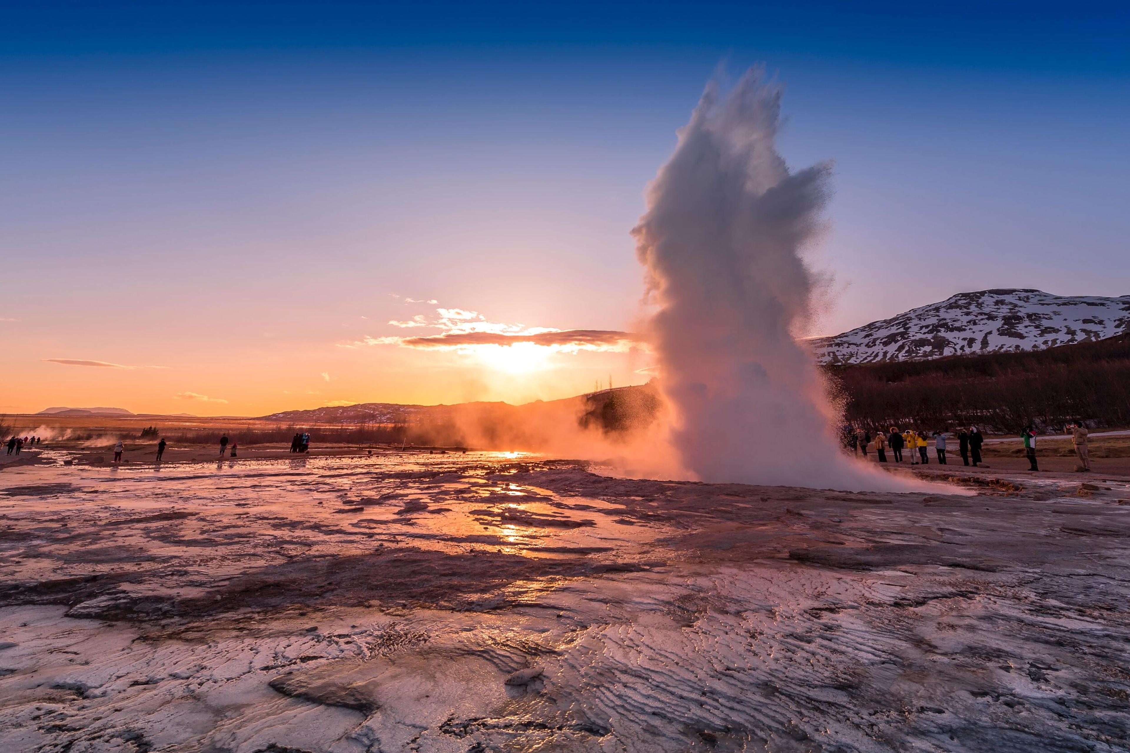 A geyser erupts with a powerful spray of water and steam at sunset, creating a dramatic scene against the orange and pink hues of the sky. People in the background observe the natural phenomenon.