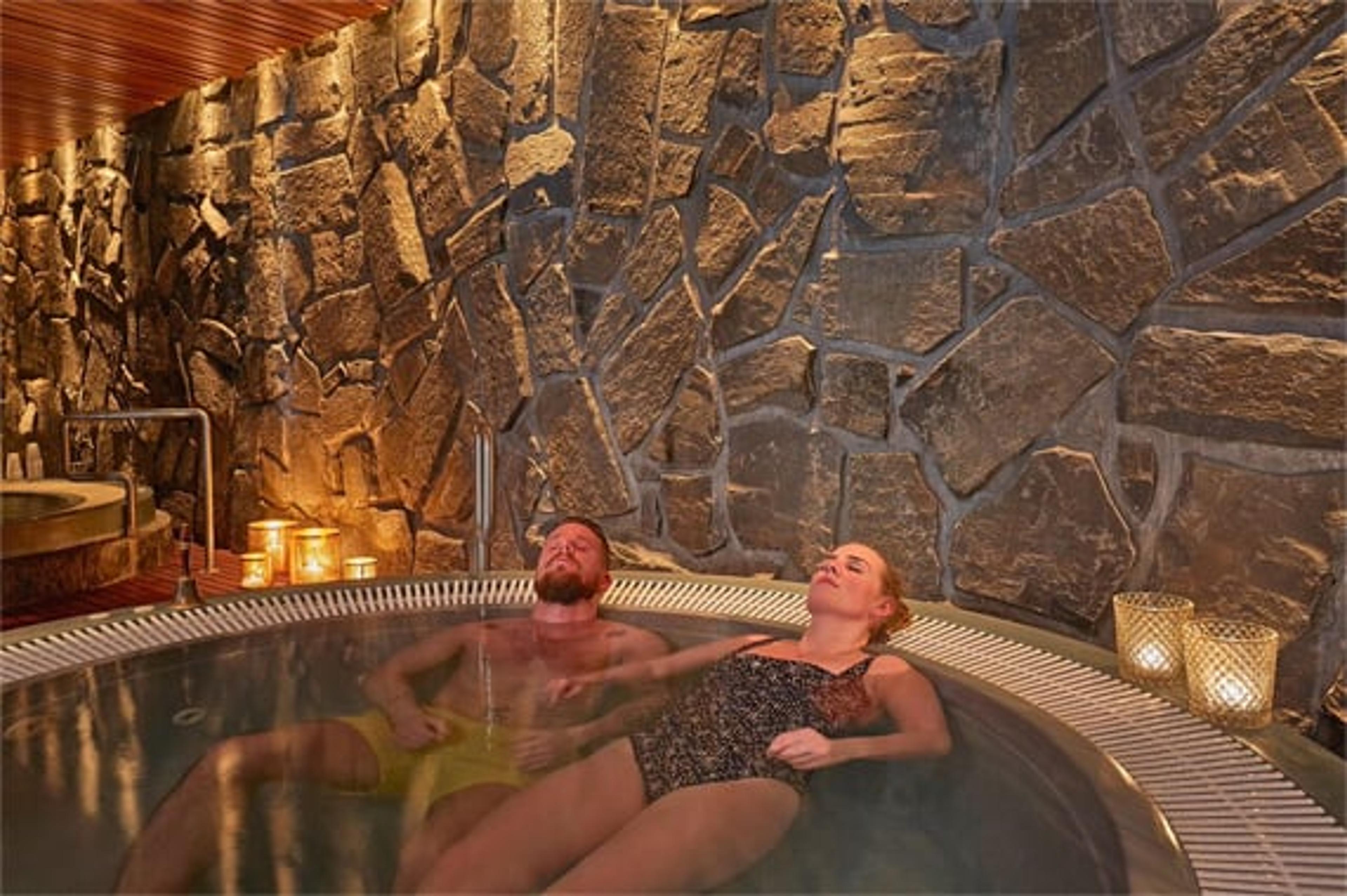 Two people relaxing in a cozy hot tub with stone walls and warm candlelit ambiance.