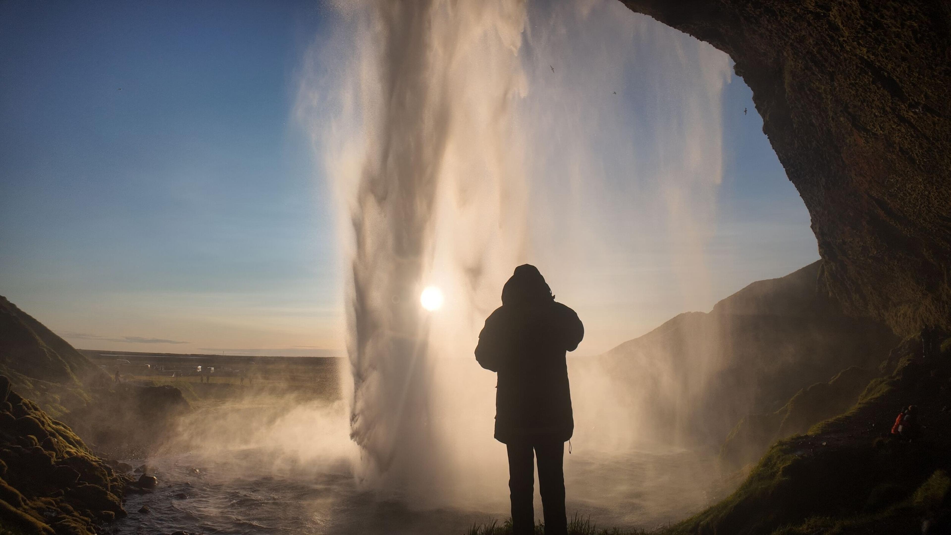 A person stands silhouetted behind a powerful, misty waterfall with the sun shining through, next to a rocky cave.