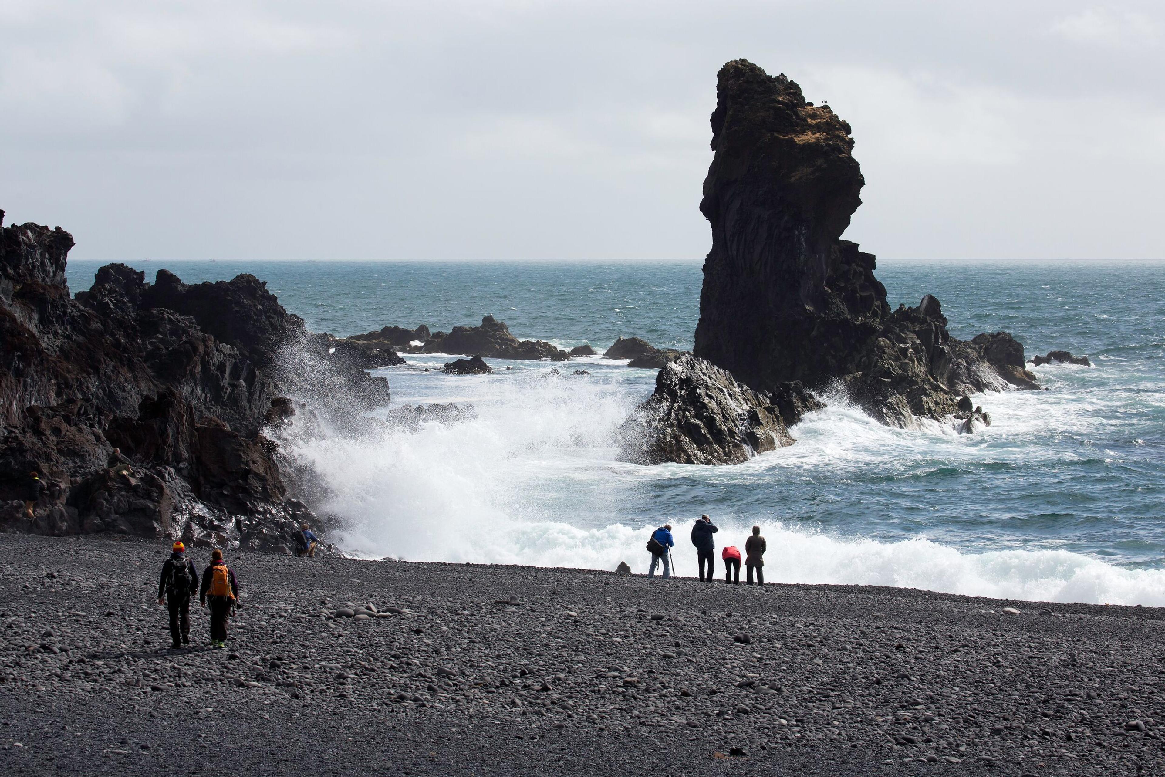 People on a black sand beach with dark sea stacks and crashing waves.