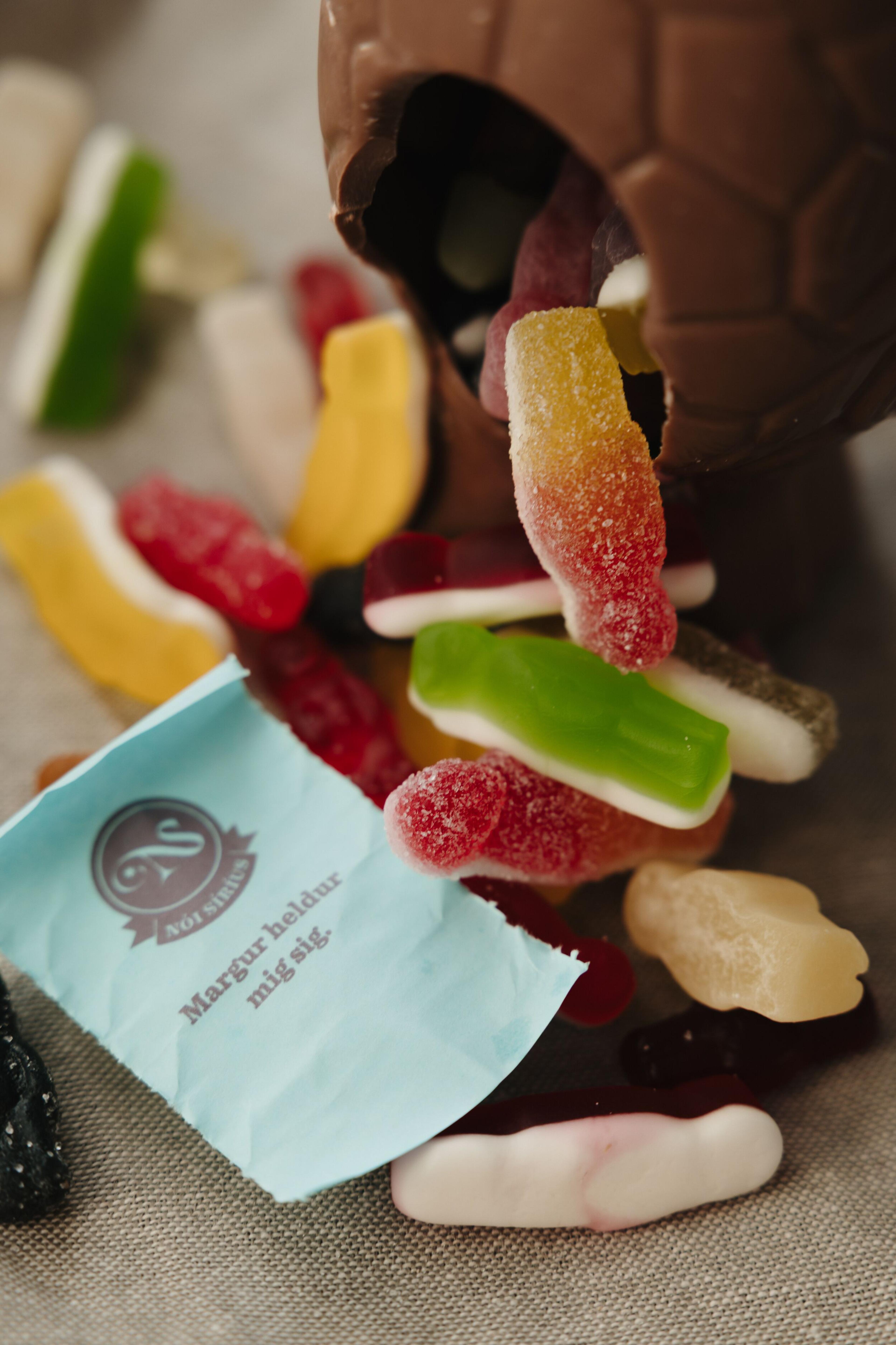 Colorful gummy candies spilling from a chocolate egg onto a light brown fabric, next to a blue candy wrapper.
