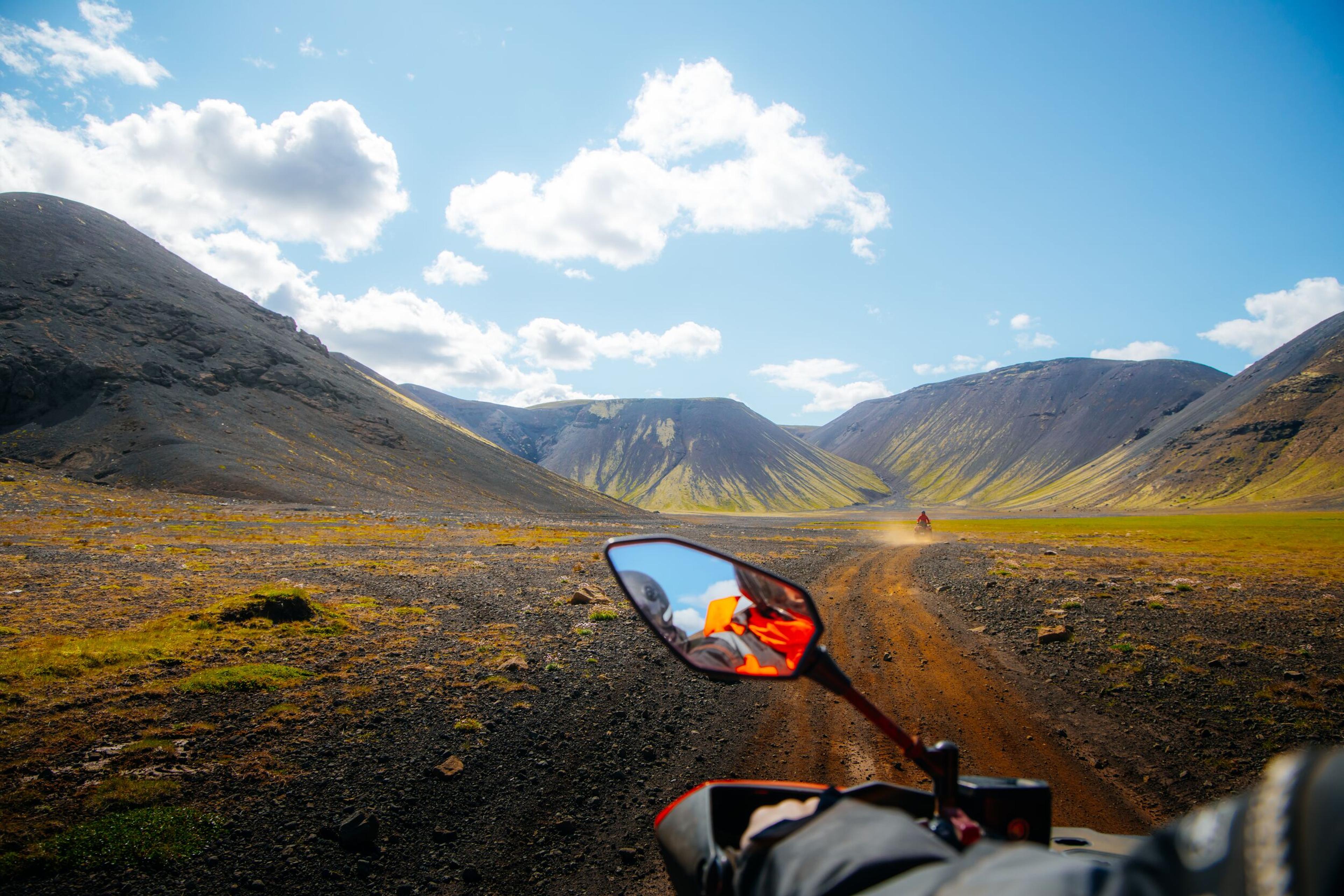 First-person view from an ATV on a dirt road through a rugged valley with mountains, another ATV visible in the distance.