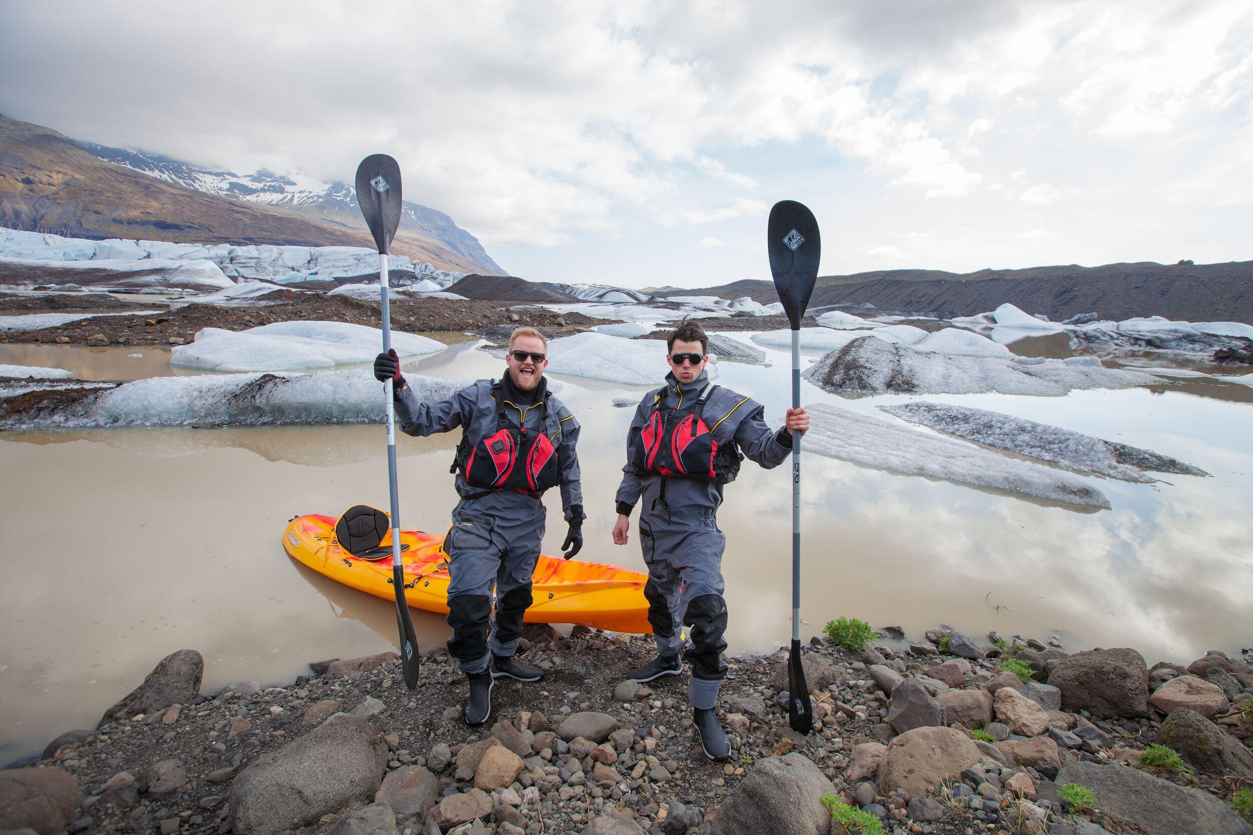 Two men in dry suits and life vests hold paddles beside an orange kayak on a rocky shore with glaciers in the background, perfect for kayaking in Iceland.