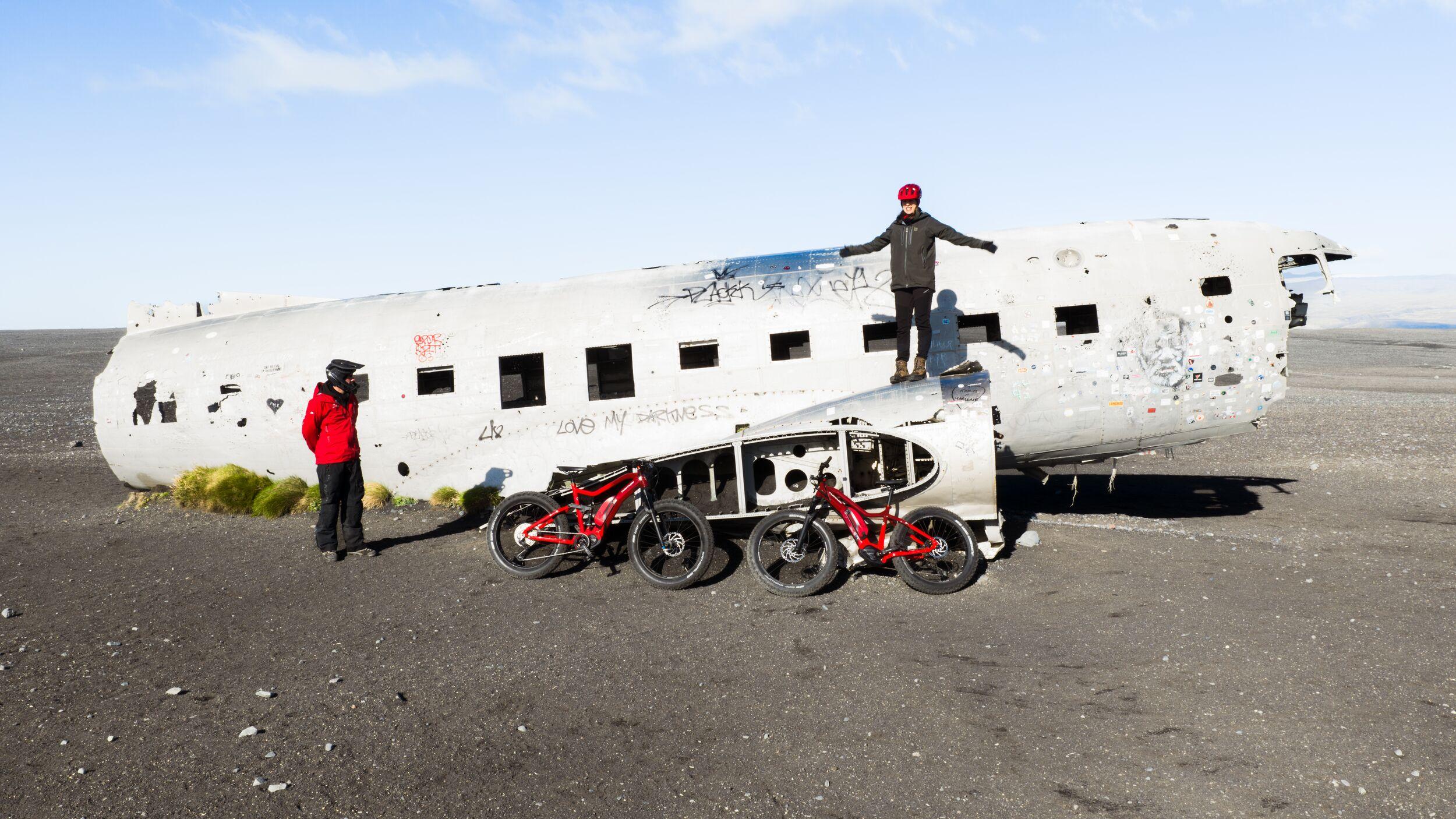 Two people at the Sólheimasandur Plane Wreck with their E-bikes (fat bikes) lying on the sand in front of the plane.