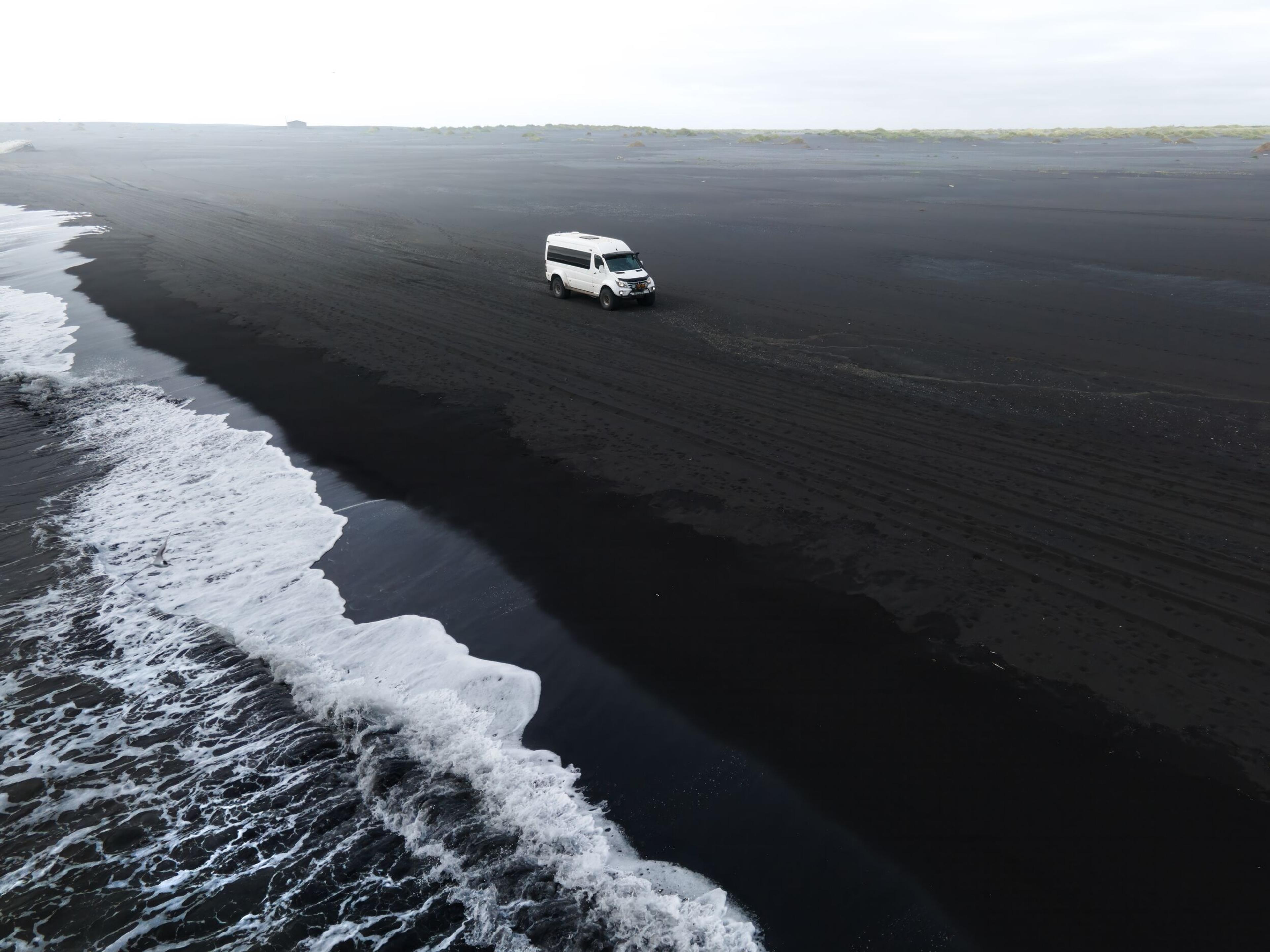 A large white monster truck making its way across a black sand beach by the shoreline.