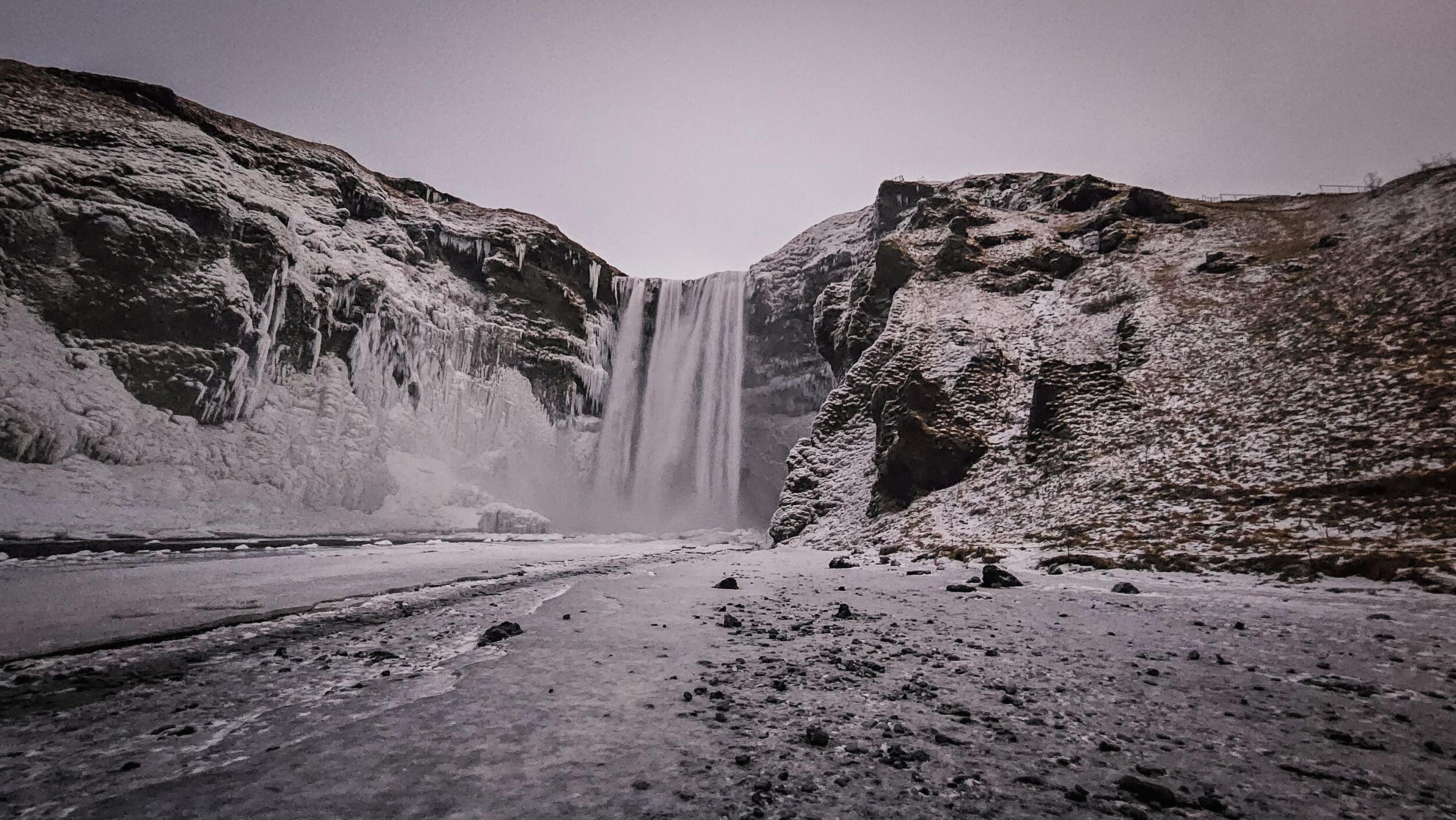 A powerful waterfall plunges between snow-covered cliffs and icicles into a frozen, rocky foreground under a grey sky.