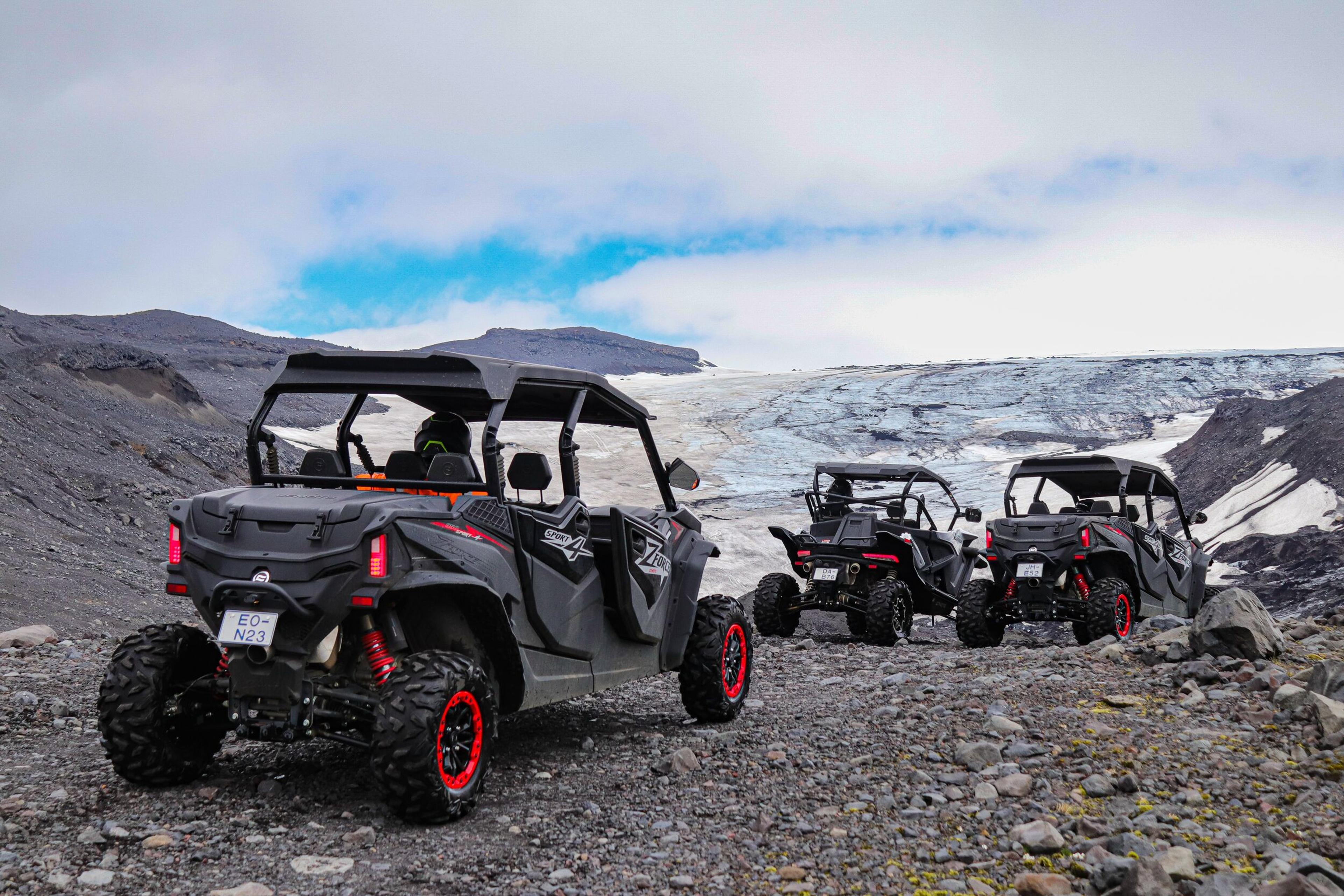 Three black ATVs with red accents driving on rocky terrain towards a glacier under a partly cloudy sky.