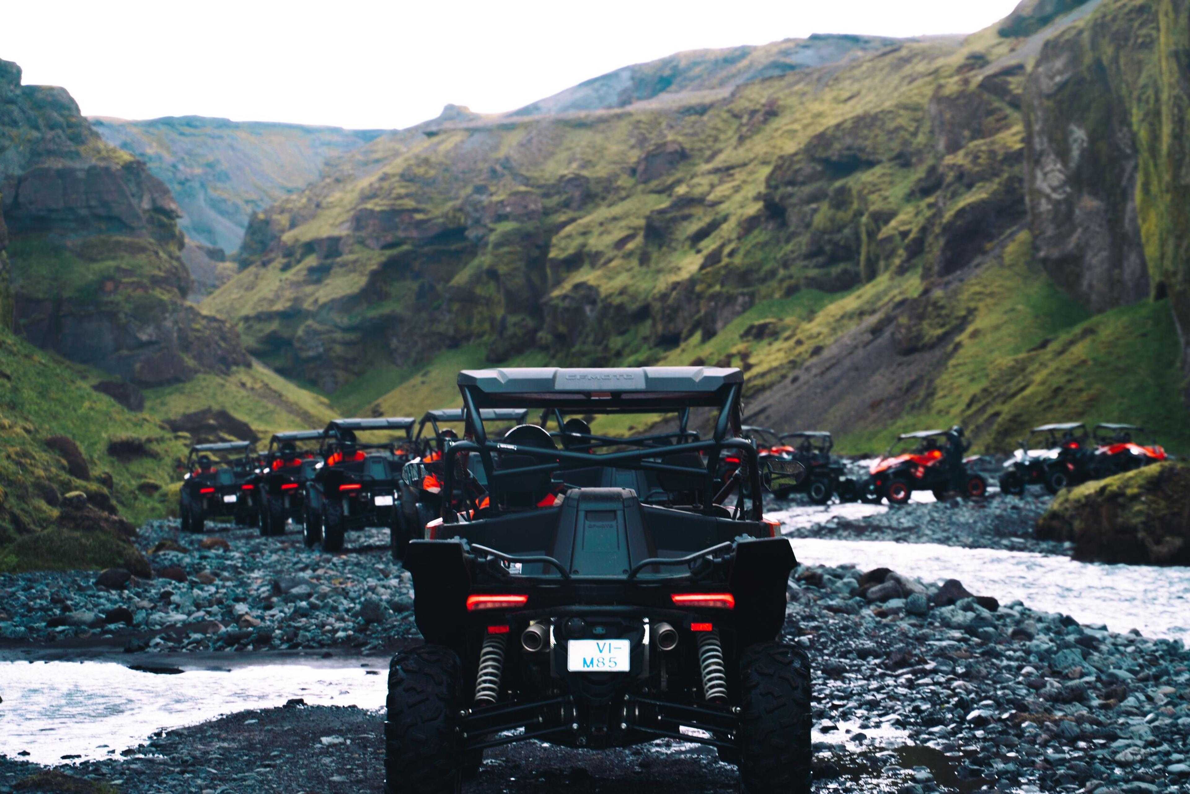 A line of UTVs driving through a rocky riverbed in a green canyon.
