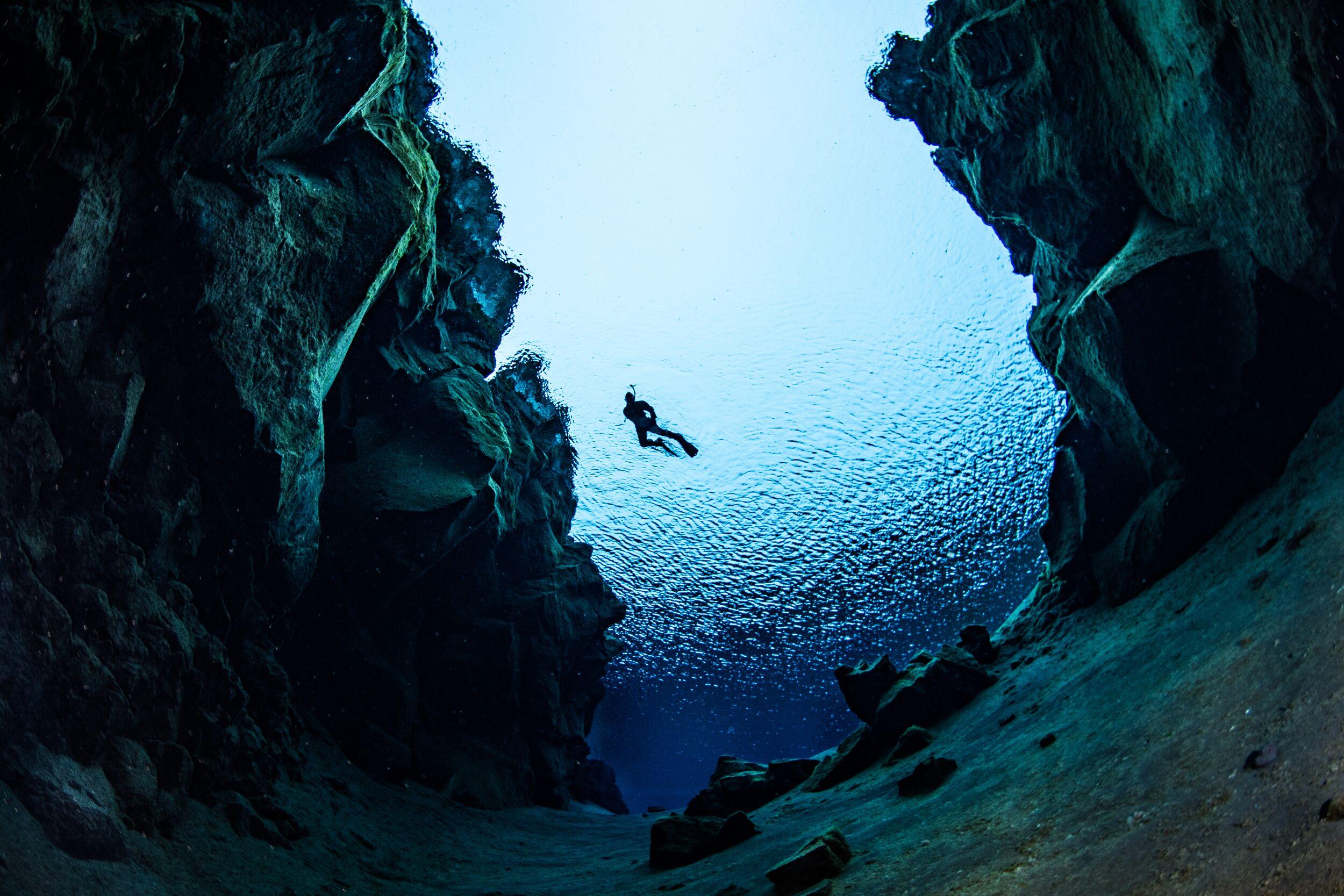 A snorkeler swims in a sunlit underwater chasm with dark rock walls.