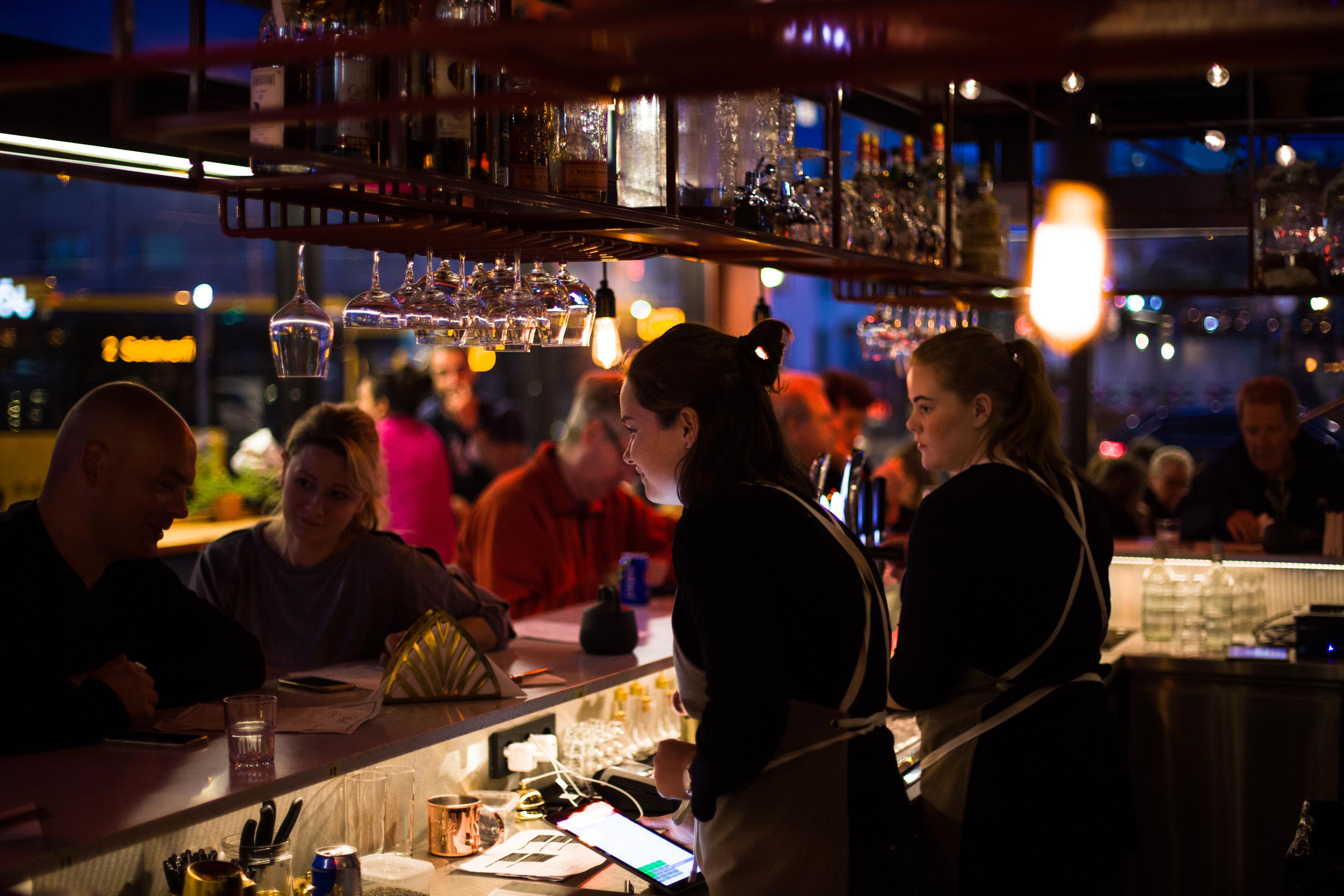 Evening scene in a Reykjavík food hall, with bartenders serving drinks to patrons at a well-lit bar counter.