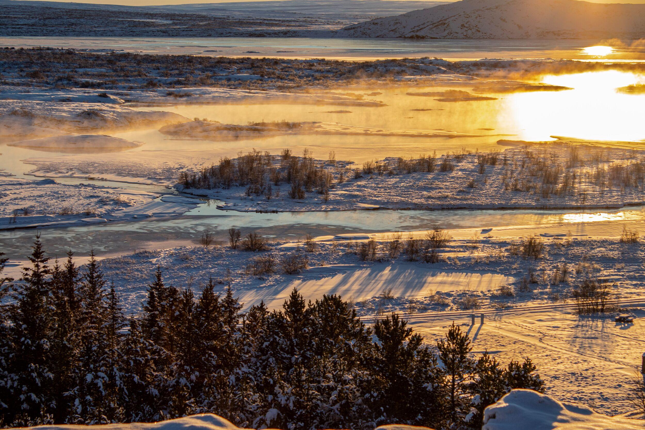 Golden sunset over a snowy river landscape with mist rising from the water.