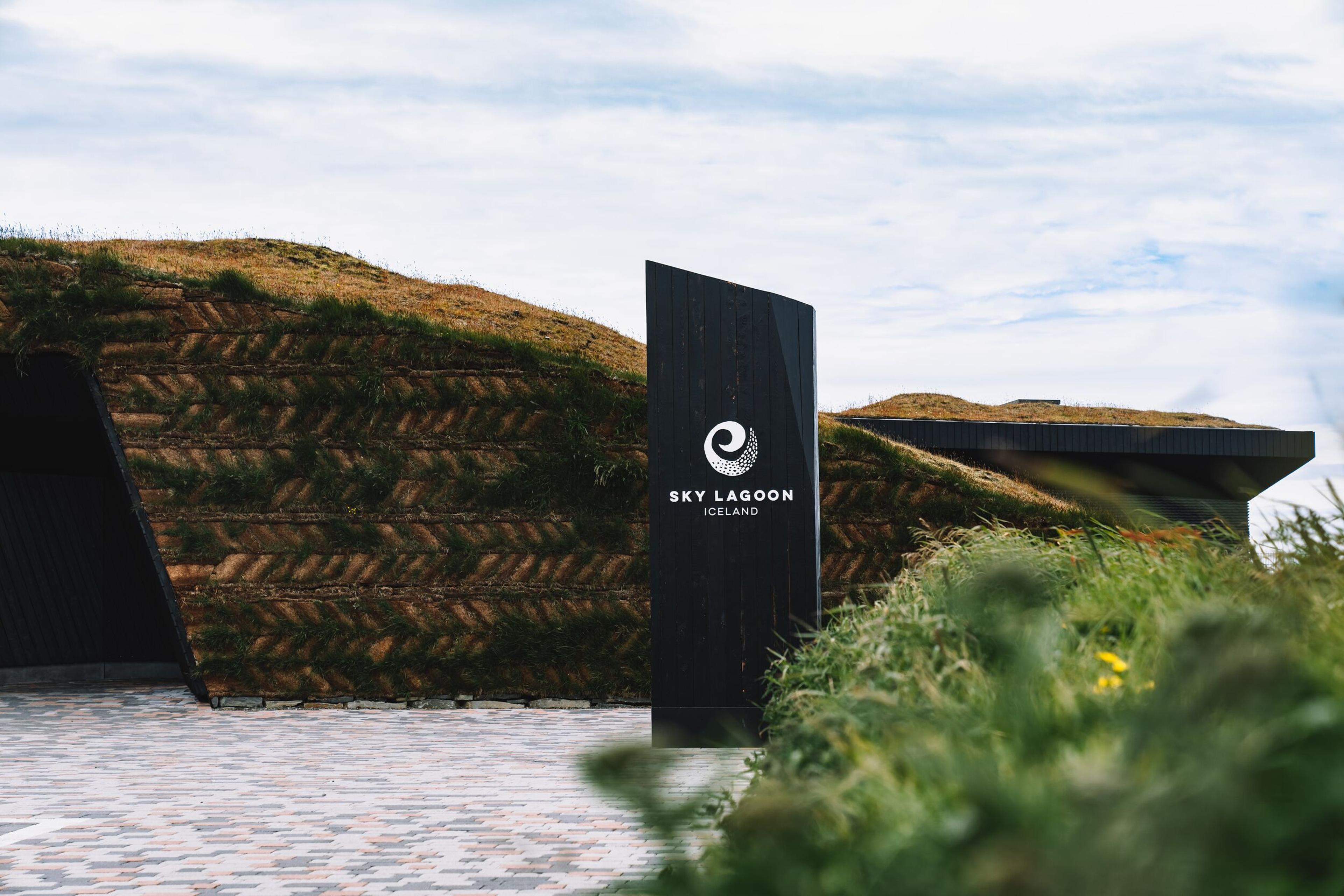  Sky Lagoon entrance with a black sign in front of a grass-covered, earth-toned building and a cobblestone walkway in Reykjavík.