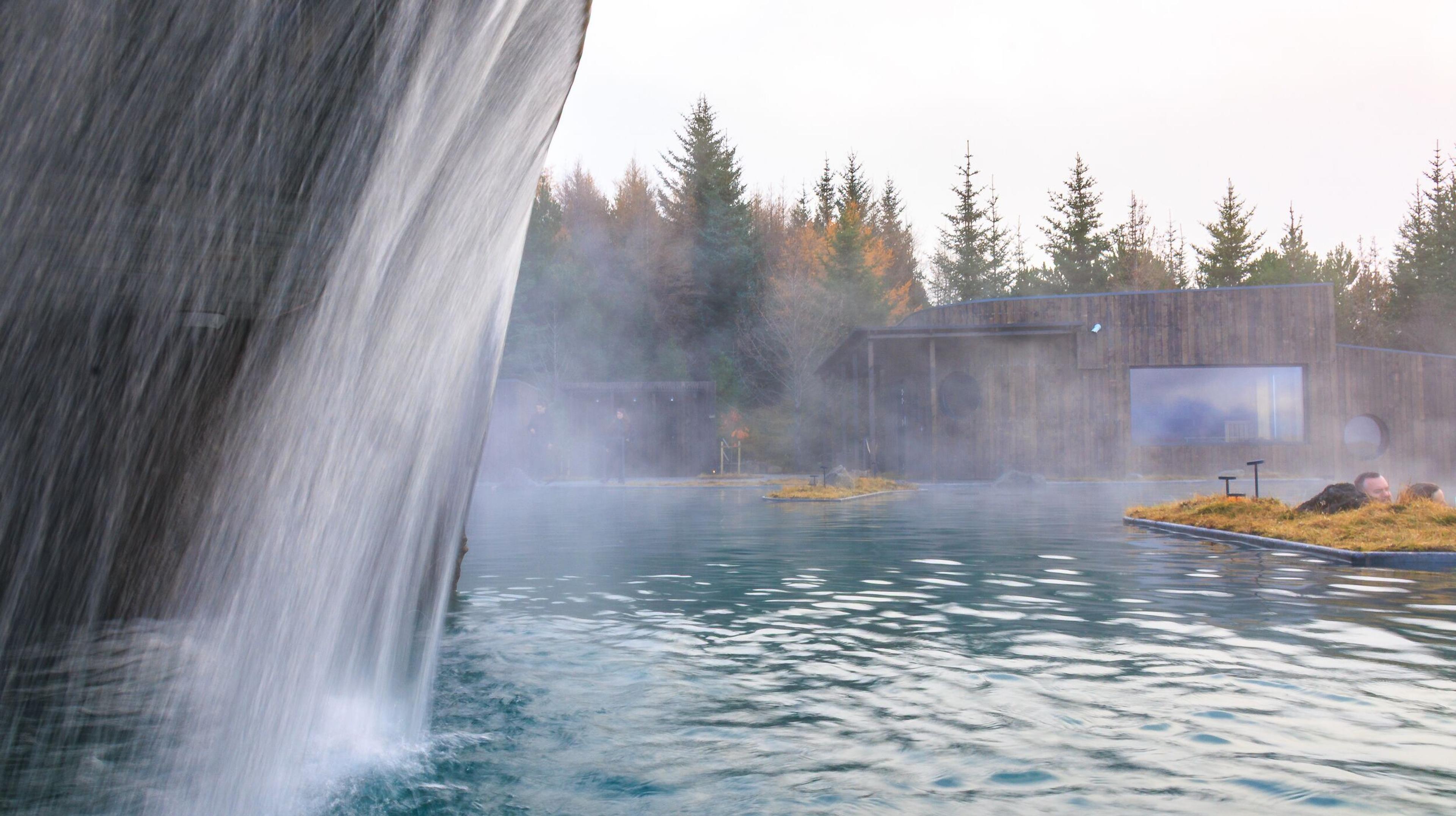 Steaming blue geothermal lagoon with a waterfall, a person on a floating mat, and a modern wooden building in the background.