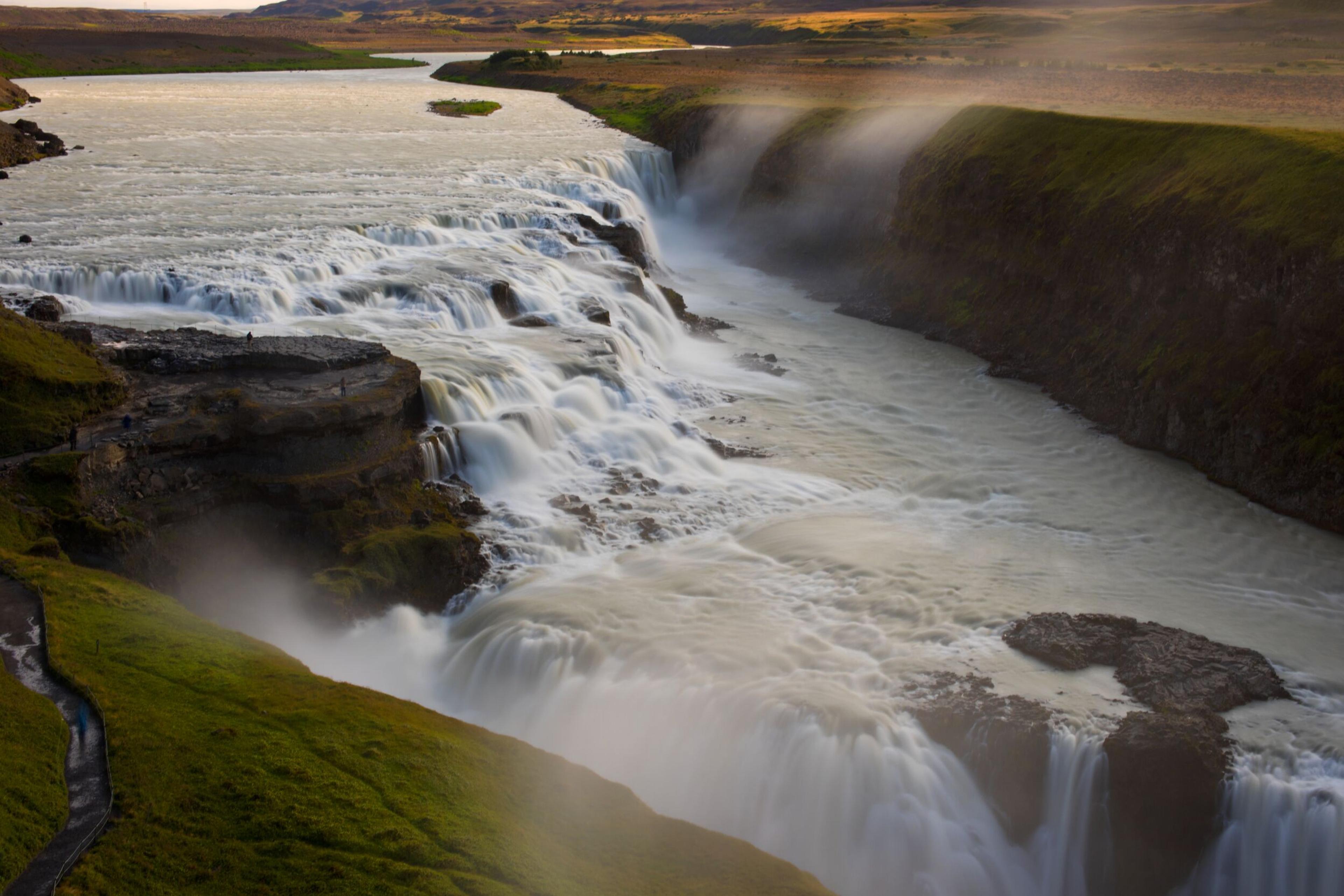 Powerful milky-white waterfalls cascade through a green canyon, with mist rising.