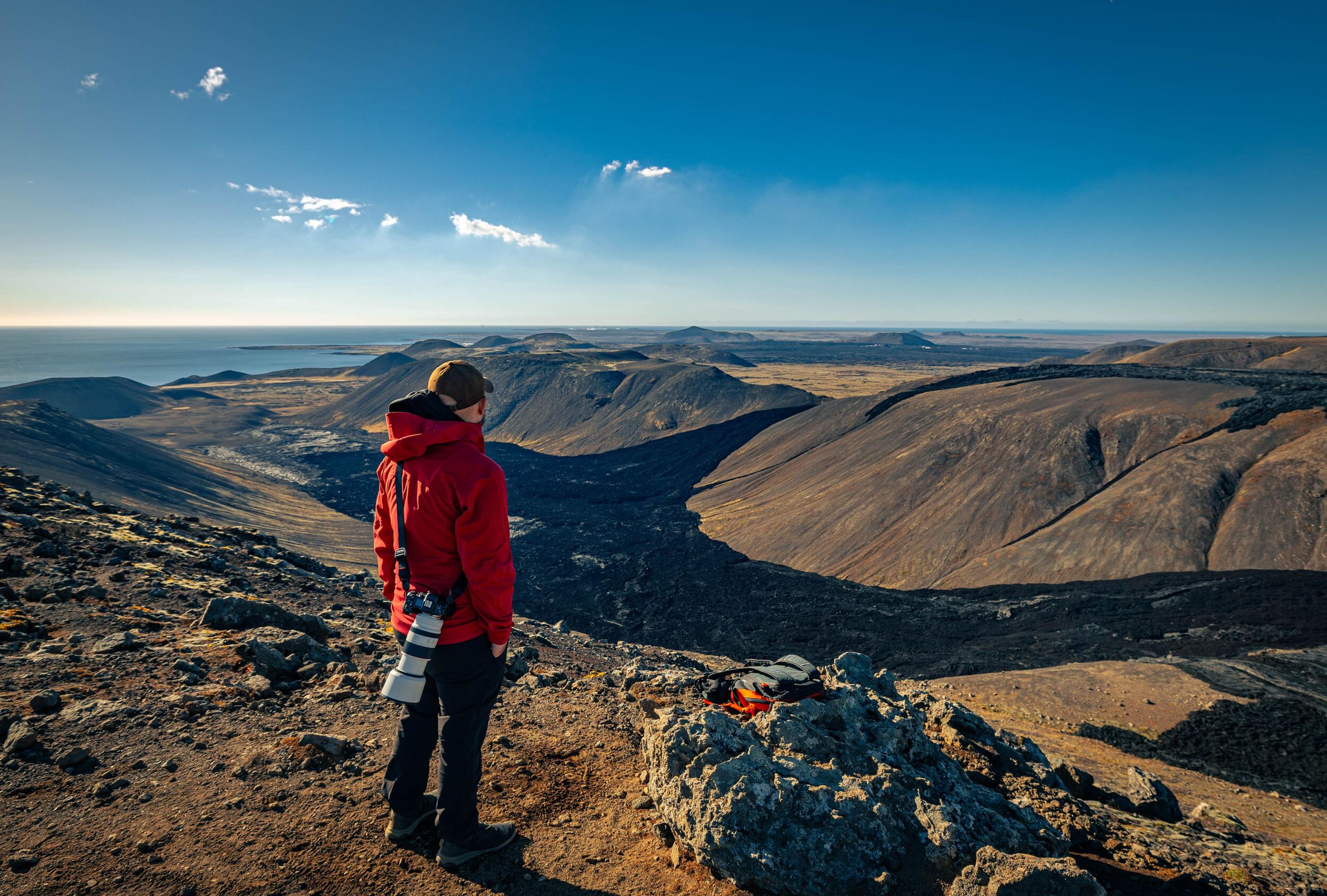 Person in red jacket stands on rocky terrain, overlooking a vast mountainous landscape with clear skies and distant horizon.