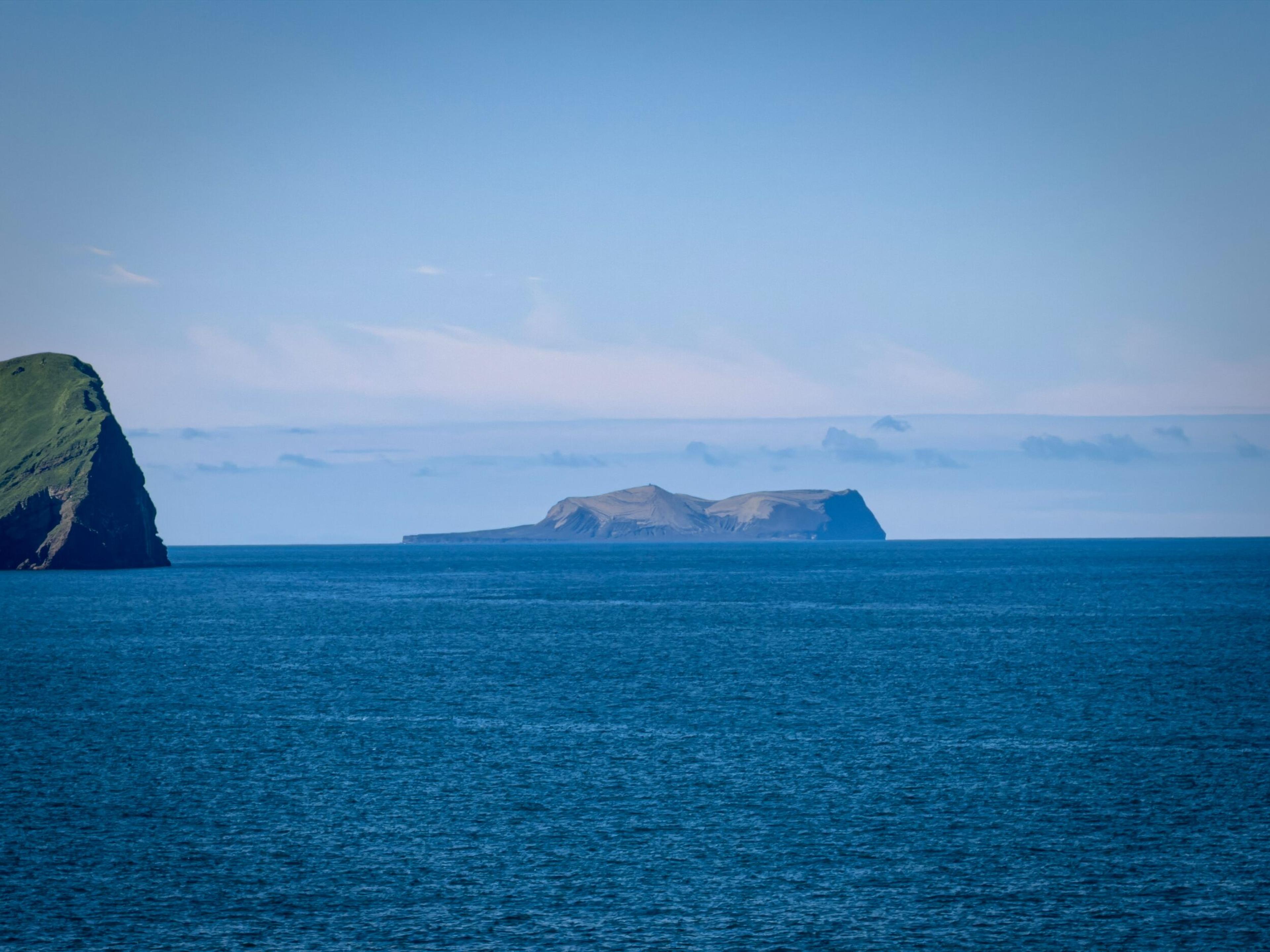 A green island cliff on the left, with another remote island in the distance across a blue ocean under a clear sky.