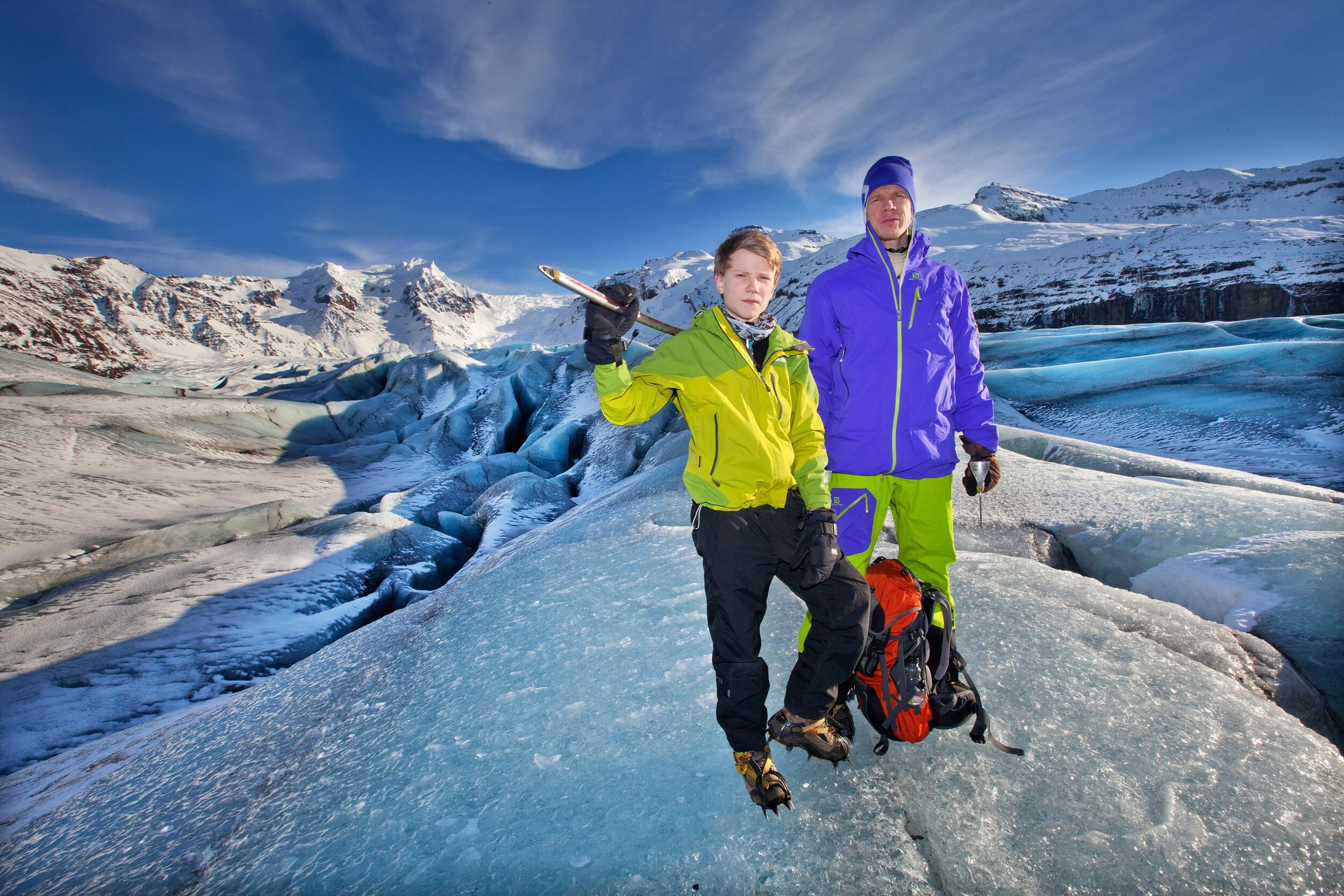 Two people in colorful winter gear stand on a glacier with ice axes, surrounded by icy terrain and snowy mountains.