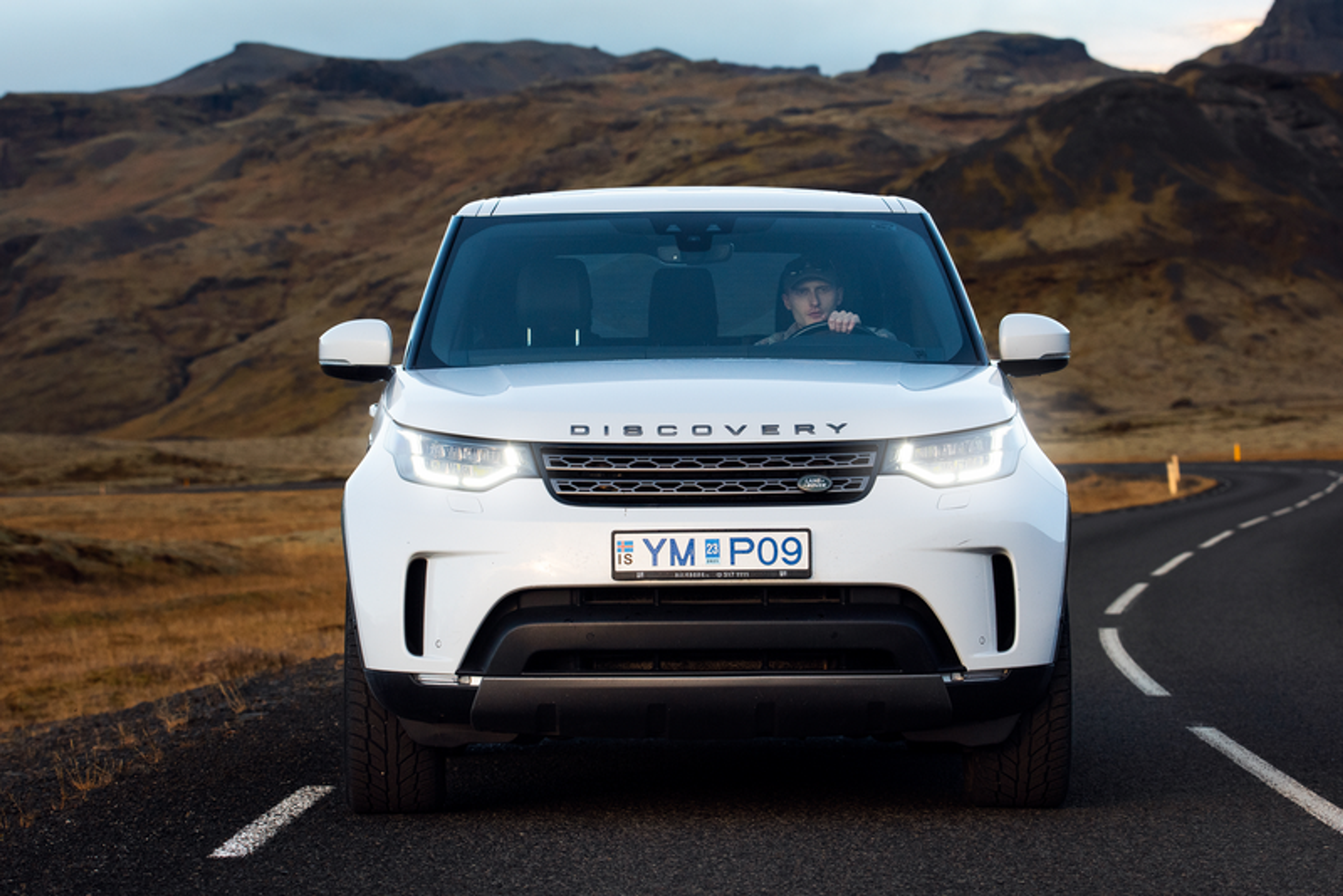 A white SUV travels along a picturesque Icelandic road with lush, rolling hills, showcasing Iceland car rental options.