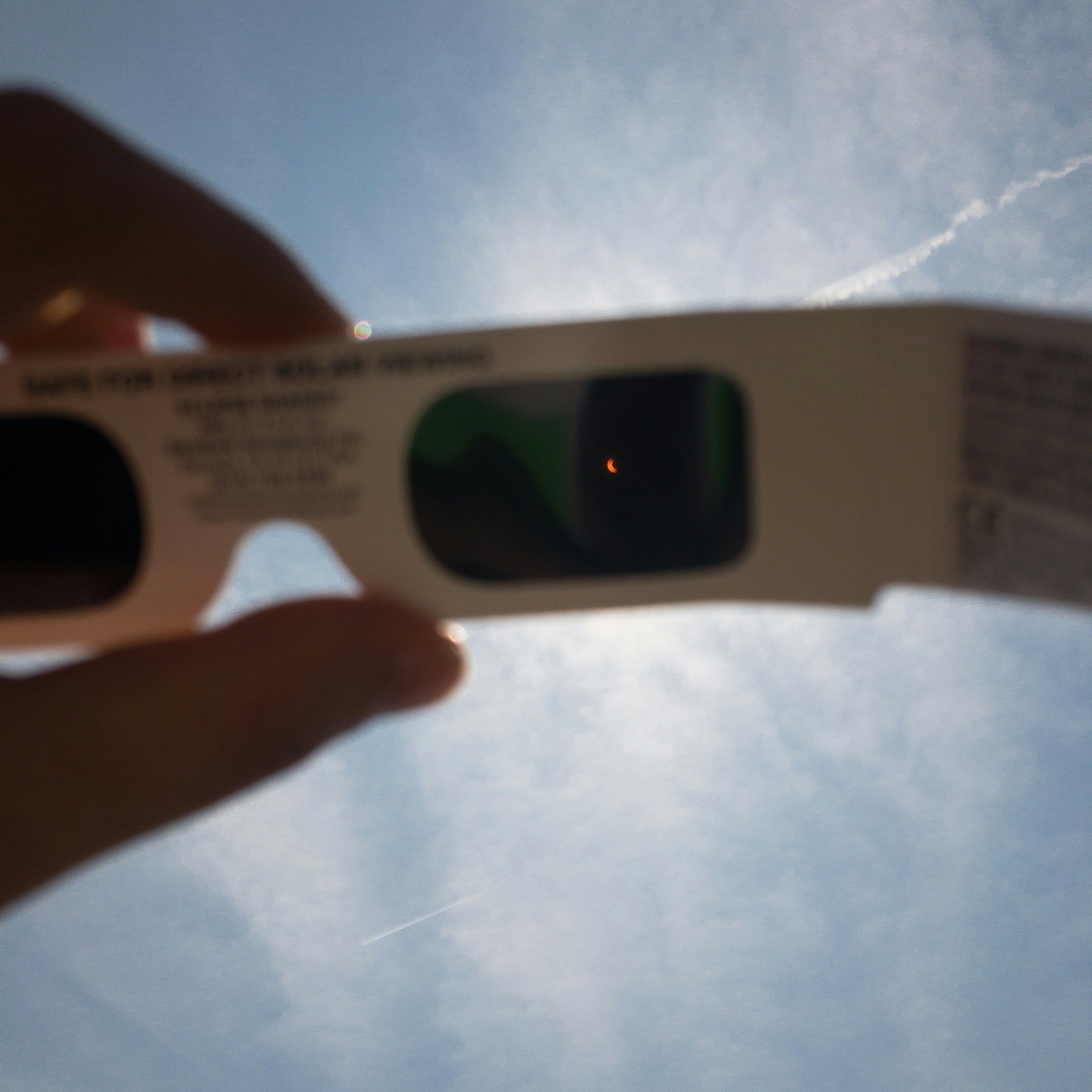 A hand holding solar eclipse glasses up to the sky, with a partially obscured sun visible through the dark lenses, ensuring safe viewing.
