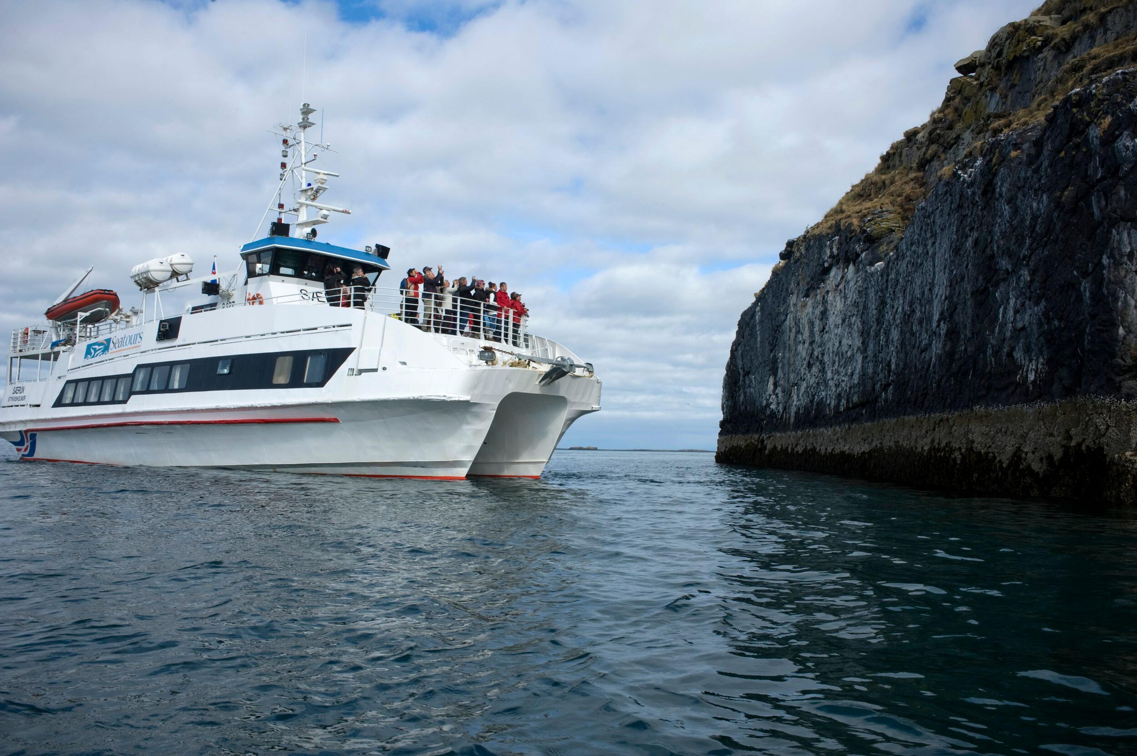 A tour boat filled with passengers navigates close to the rugged cliffs of a narrow channel, reflecting the clear waters below.
