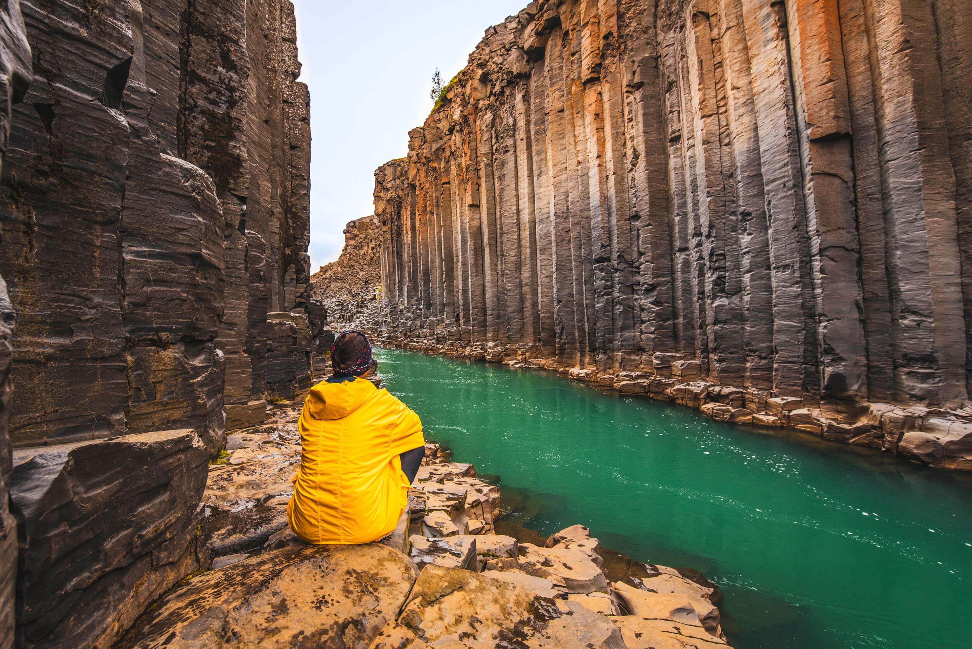 Person in a yellow jacket sitting on a rock ledge, gazing at the emerald green waters of Stuðlagil Canyon in Iceland, surrounded by distinctive tall basalt columns