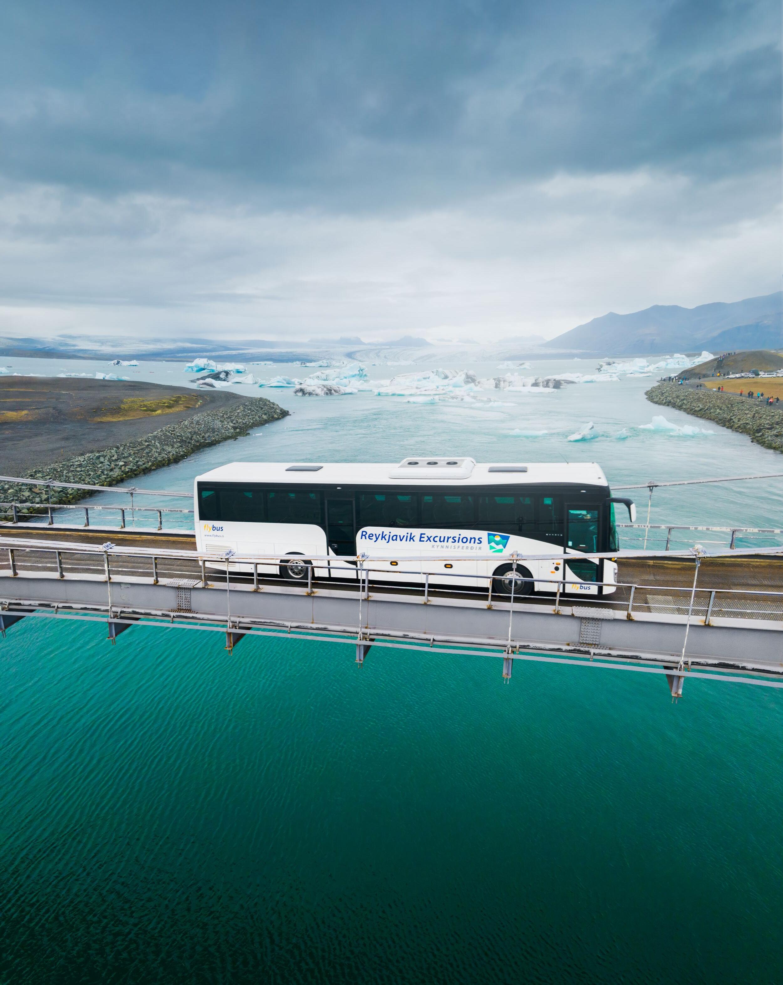 A tour bus from Reykjavik Excursions crossing a bridge with stunning views of Icelandic glaciers and icebergs in the background.