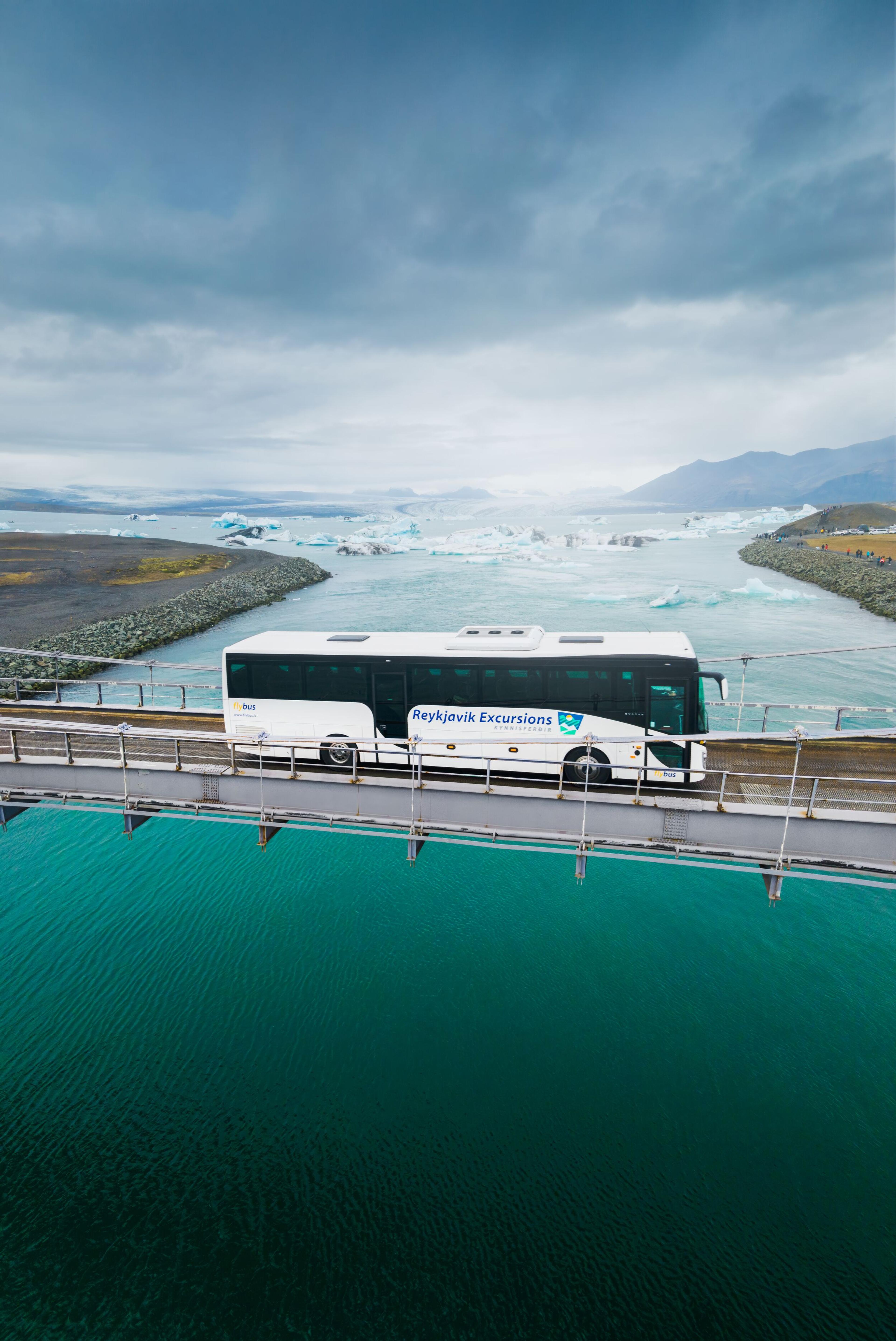 A tour bus crossing a bridge over turquoise waters with a view of a glacier and floating icebergs in the background, under an overcast sky.