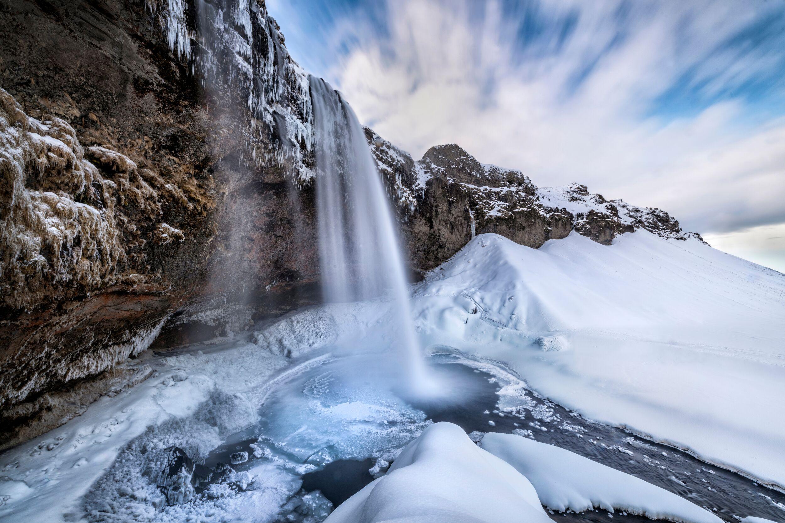 Winter waterfall plunging into an icy pool, surrounded by snow-covered cliffs under a streaky blue sky.