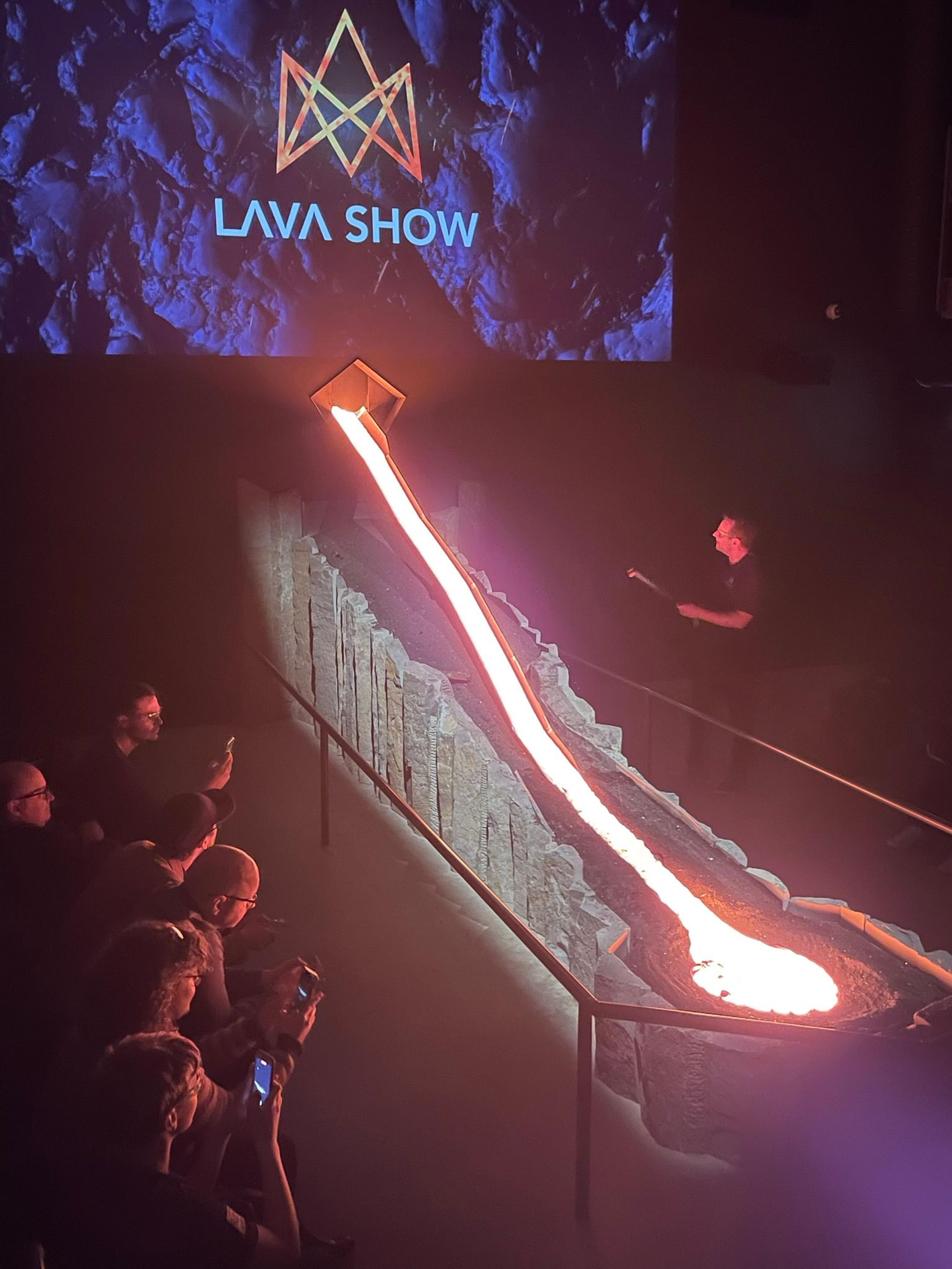 A "LAVA SHOW" with molten lava flowing down a rocky channel past an audience and a presenter.