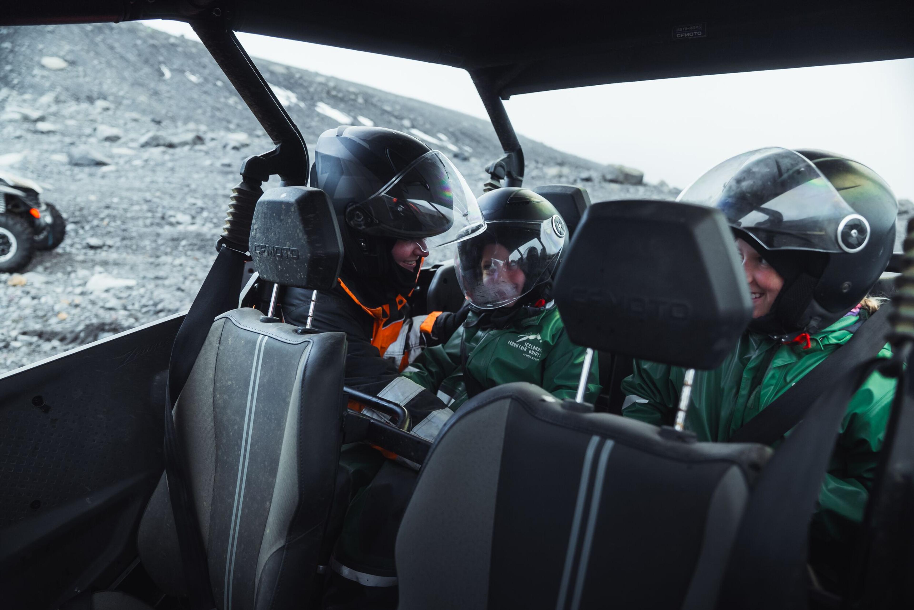 Four people in helmets smiling inside a UTV on a rocky landscape.
