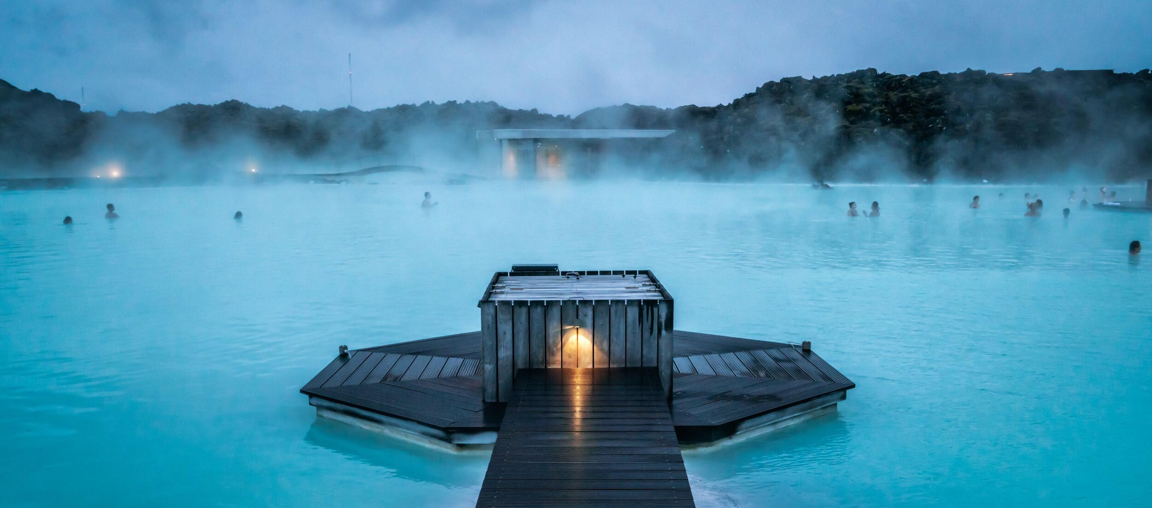 A steaming, milky blue geothermal lagoon with bathers and a lighted wooden walkway.
