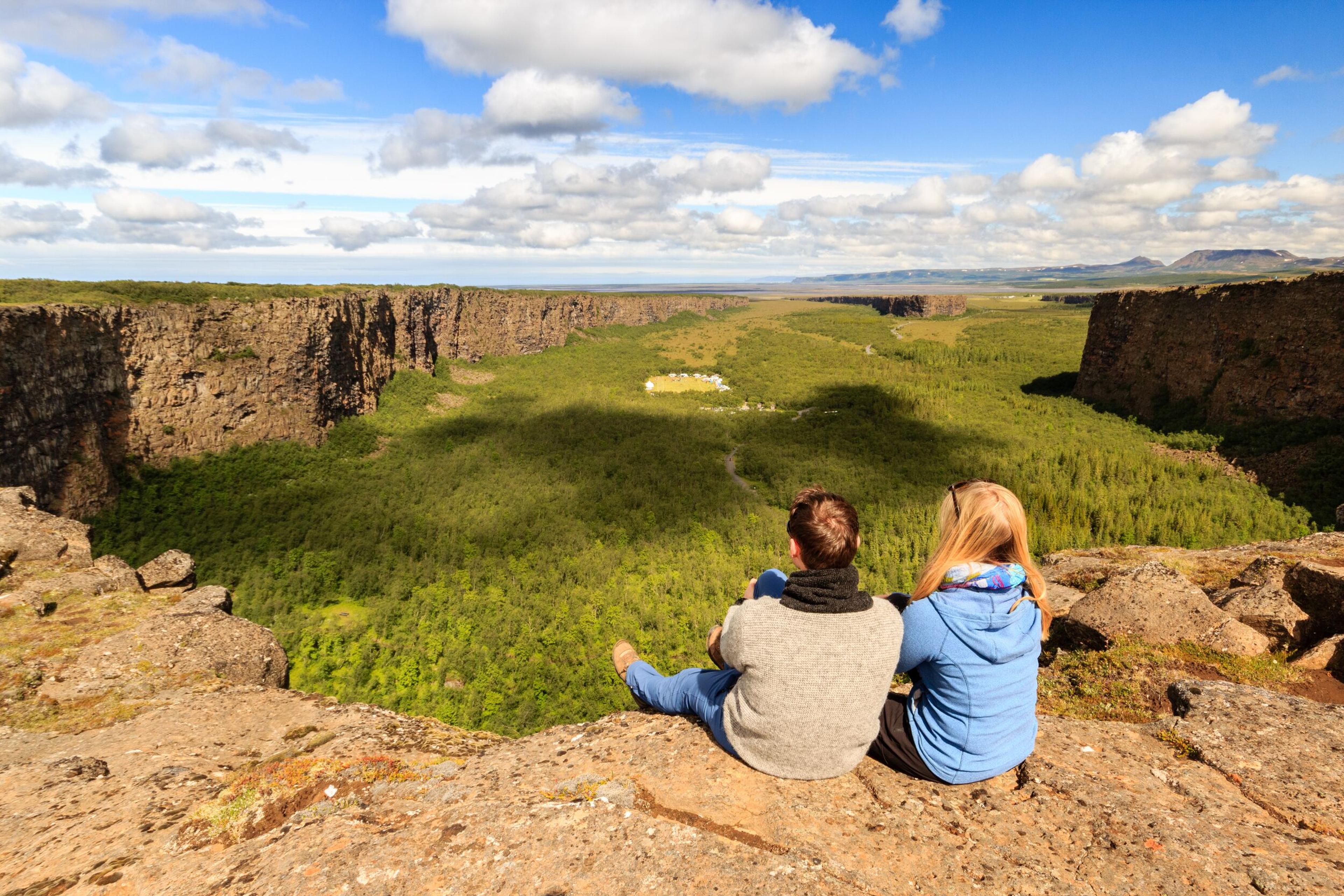 A couple sitting on the edge of a cliff overlooking the lush, horseshoe-shaped Ásbyrgi Canyon in Iceland, with dense green vegetation below and a clear blue sky above.