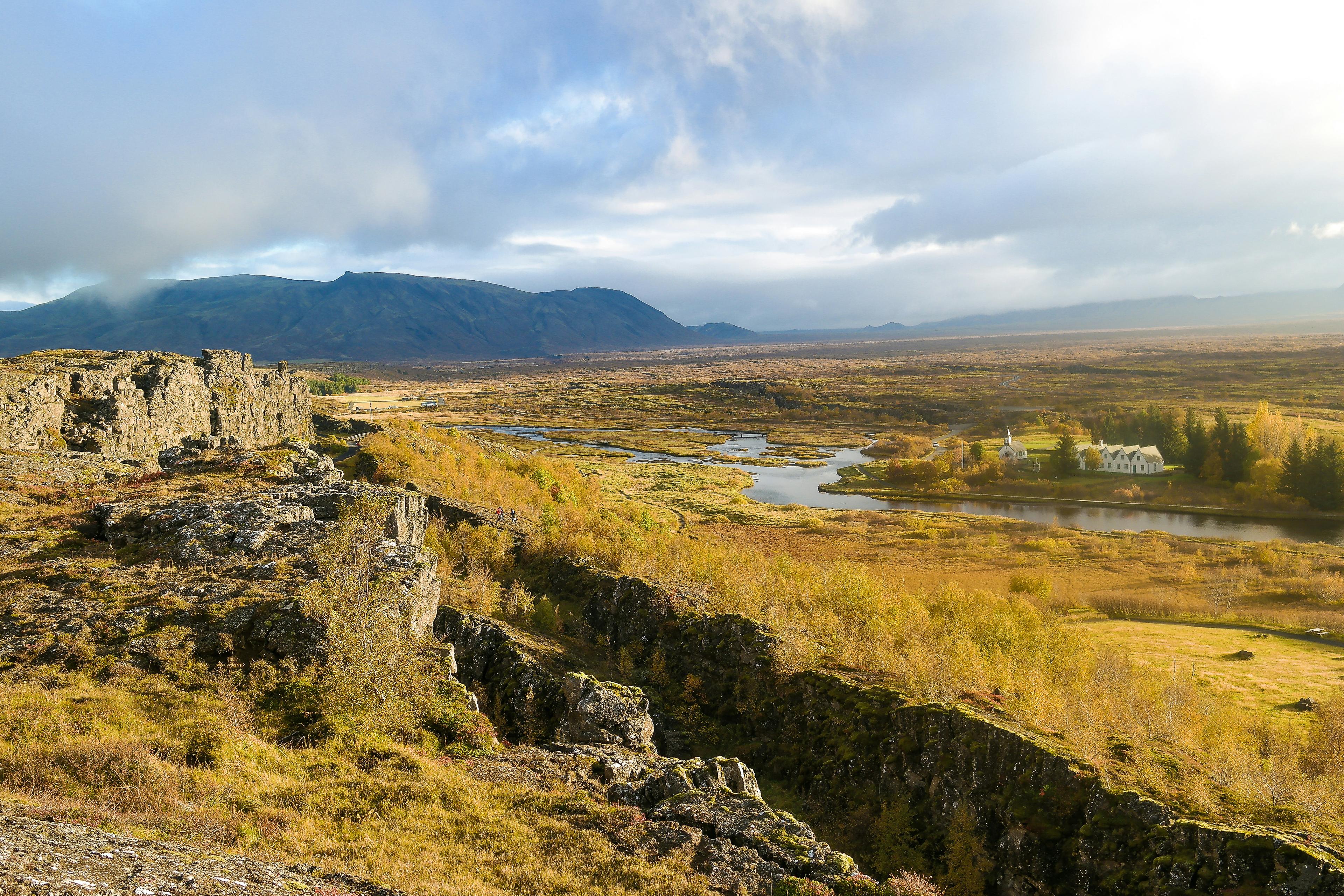 Serene landscape of Þingvellir National Park featuring a traditional Icelandic church surrounded by trees with golden foliage, reflecting in the calm waters of the Oxara River under a clear blue sky.