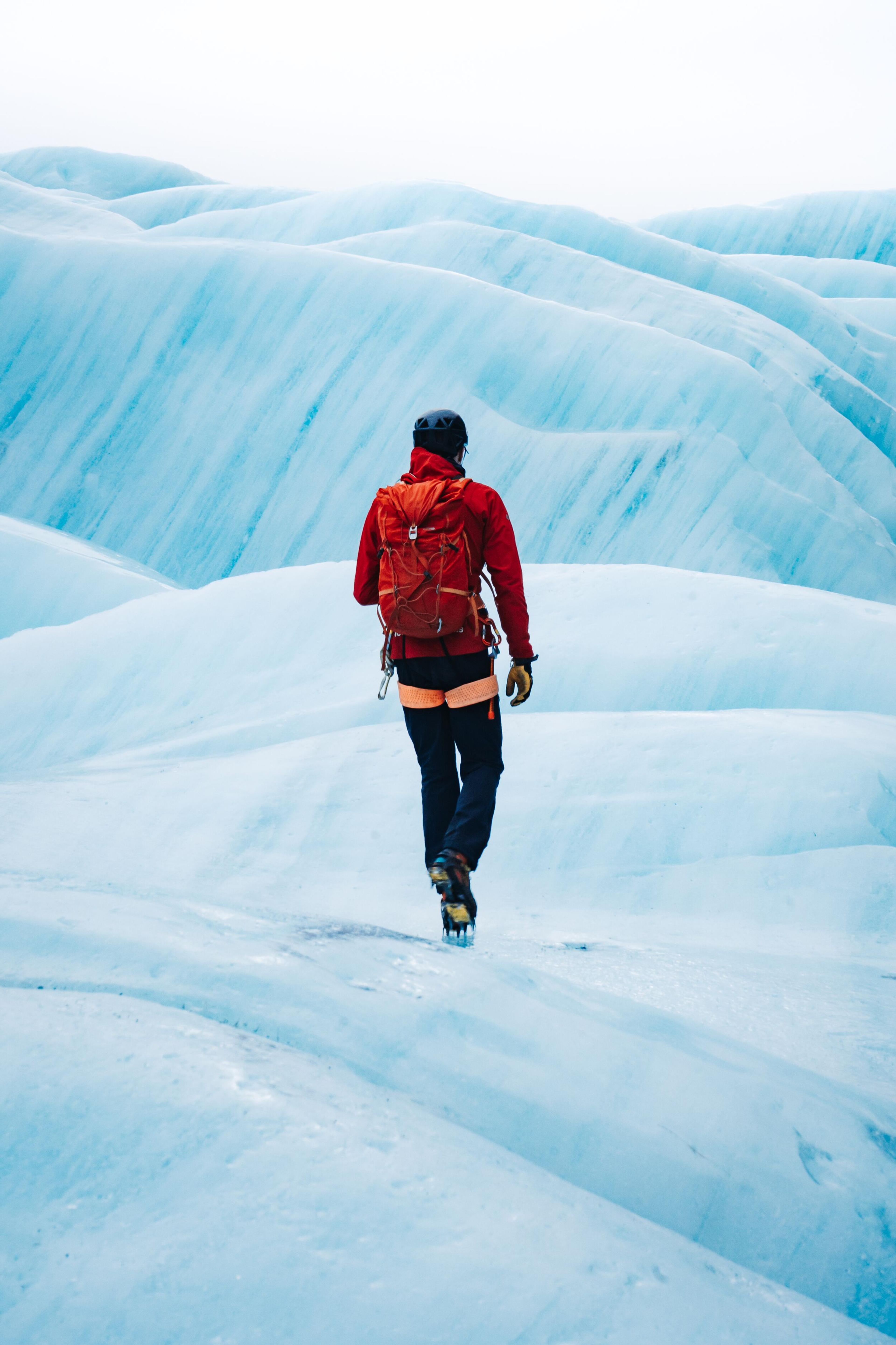 A person in a red jacket and backpack trekking on a blue glacier, moving towards an imposing ice wall, with their back to the camera