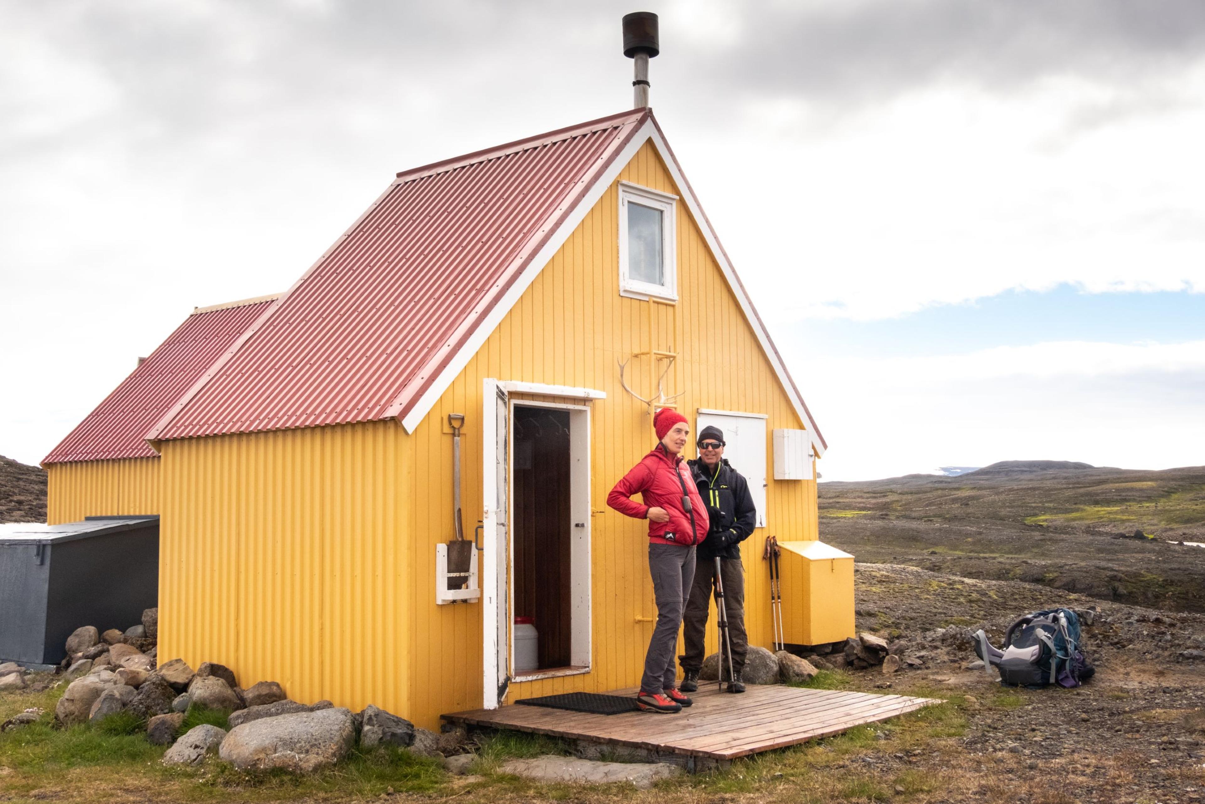 Two hikers posing in front of a quaint yellow hut in a wilderness setting.