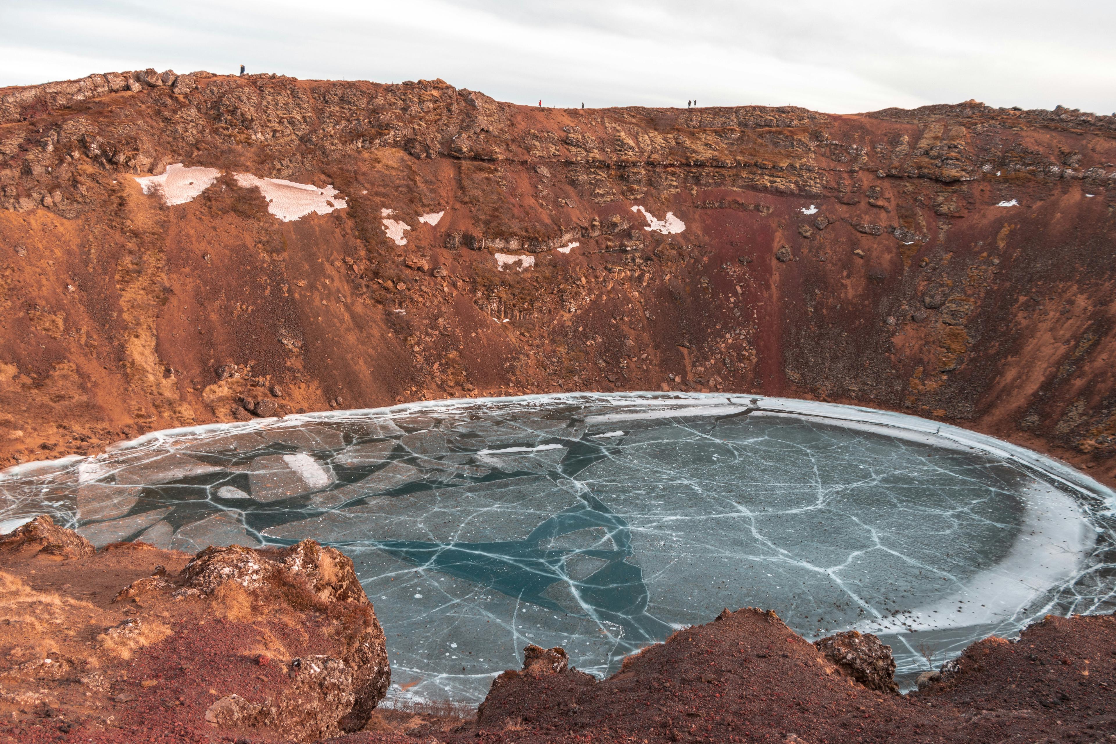 An icy crater lake with a web of cracks on its frozen surface, enclosed by rugged red cliffs, a striking contrast to the sprinkling of snow, under a soft, glowing sky.
