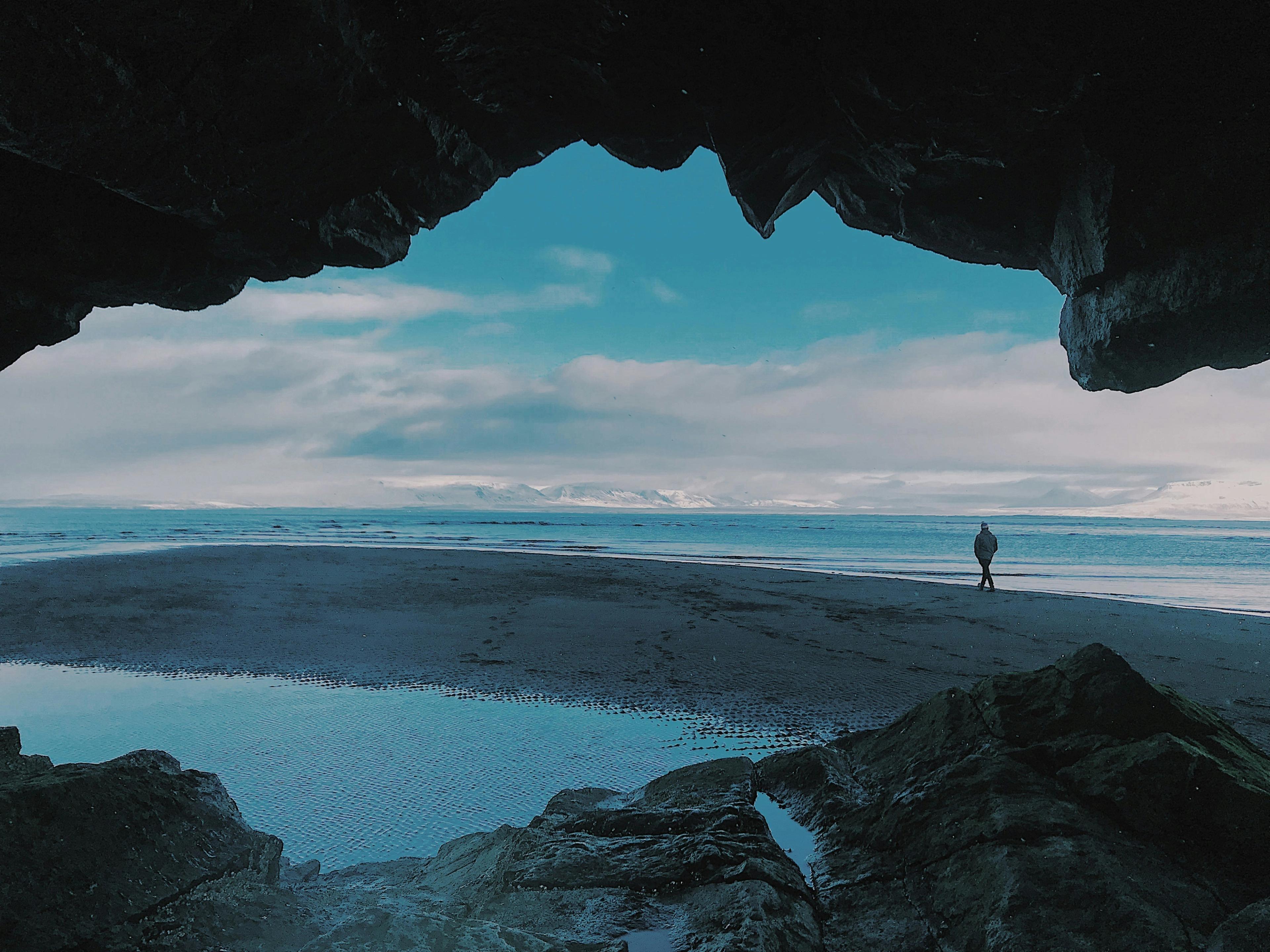View from inside a dark cave looking out at a person walking on a wide beach with the ocean, distant snow-capped mountains, and a blue sky.