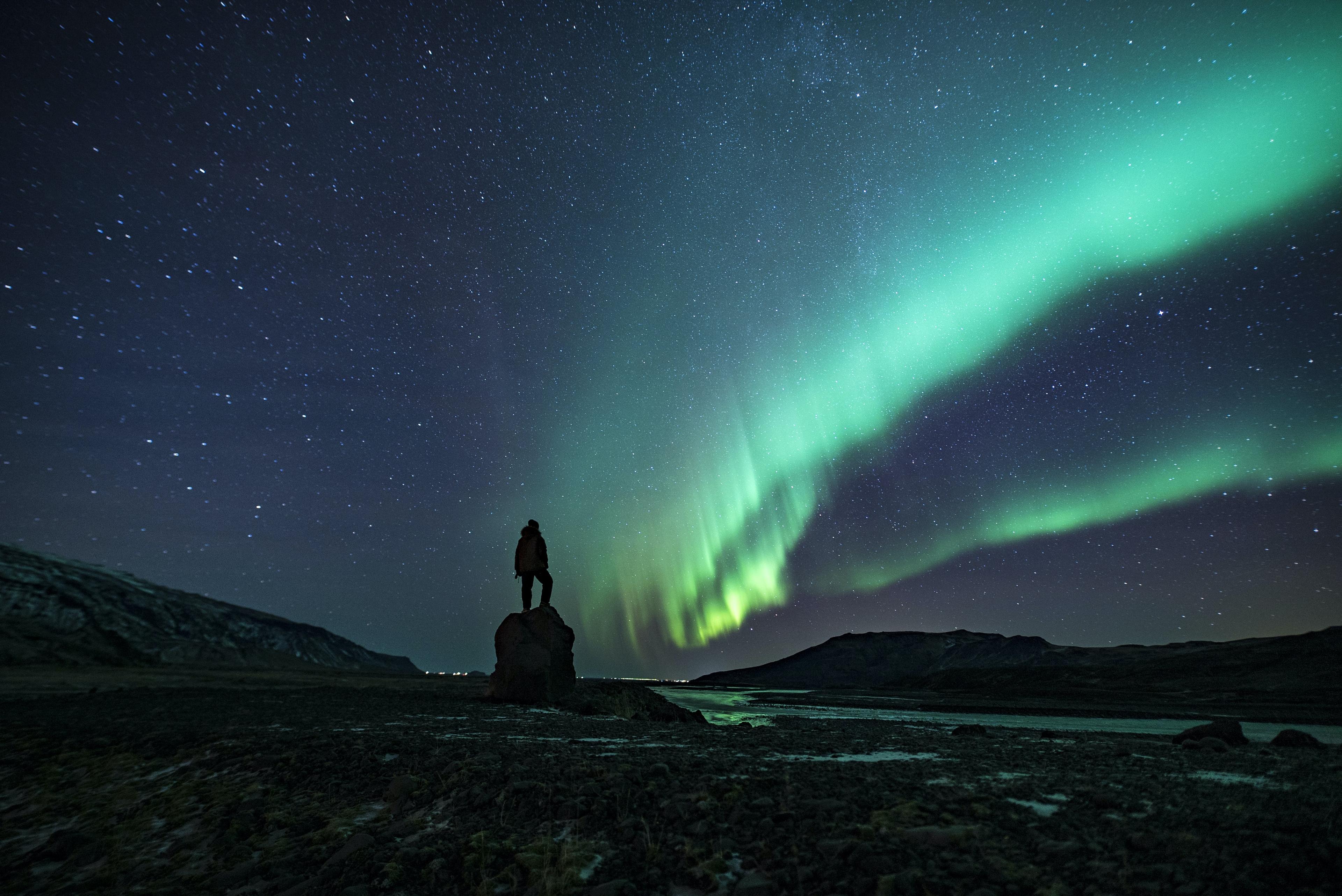 A person standing atop a rock, gazing at the Northern Lights under a star-filled sky.