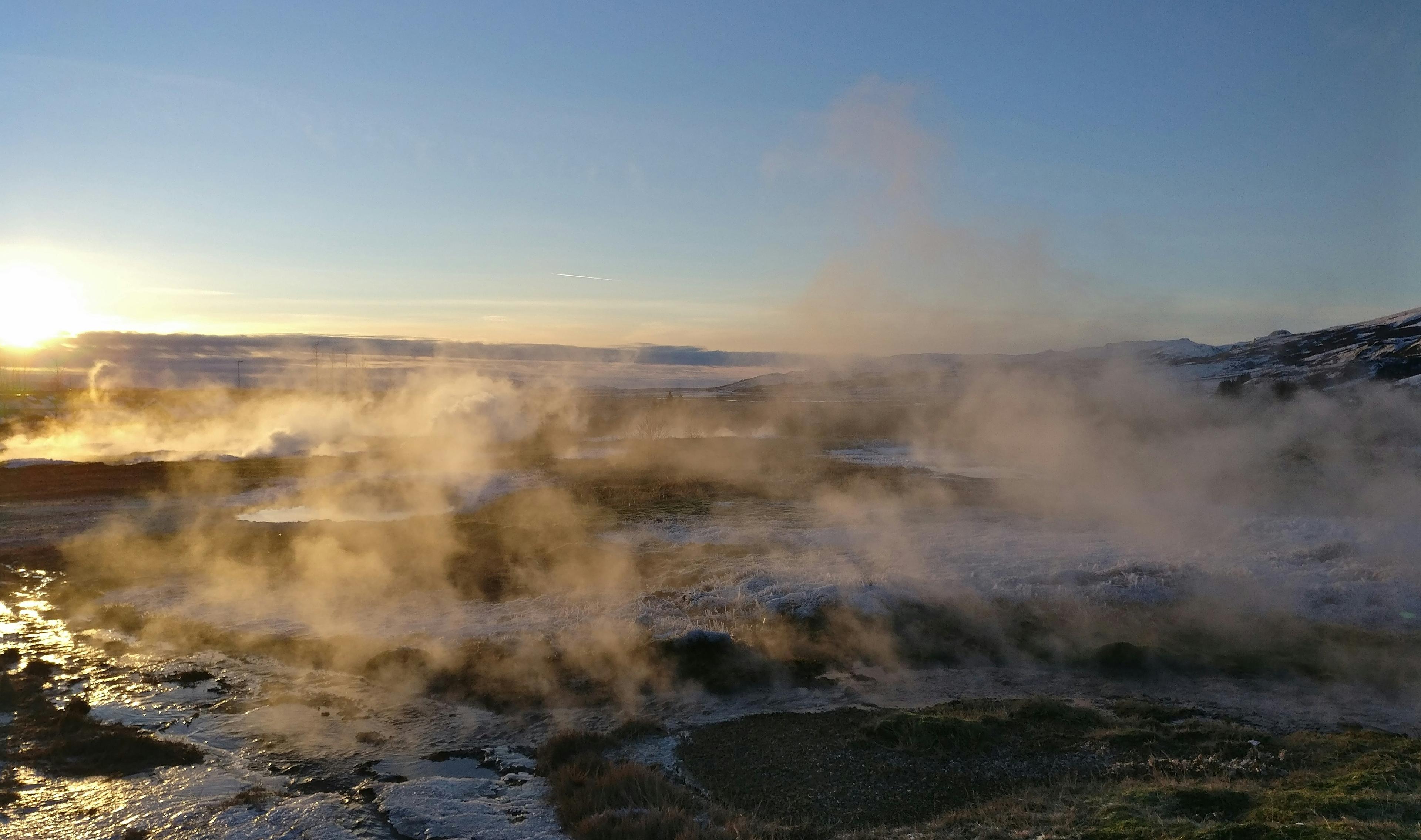 A steamy geothermal landscape captured at sunrise or sunset, with the warm glow of the sun highlighting the steam and rugged terrain, evoking the geothermal activity of areas like those found in Iceland.