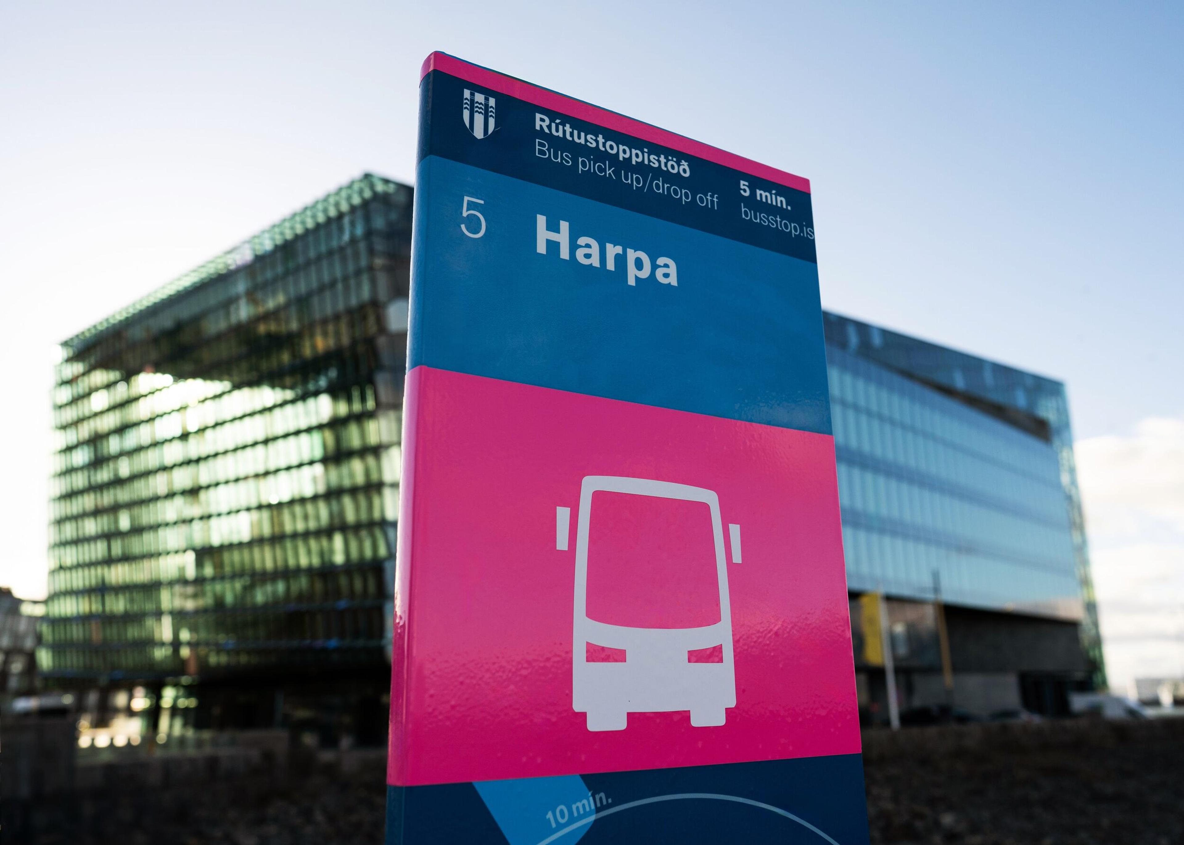 A blue and pink bus stop sign for 'Harpa' featuring a bus icon, with a modern glass building in the background.