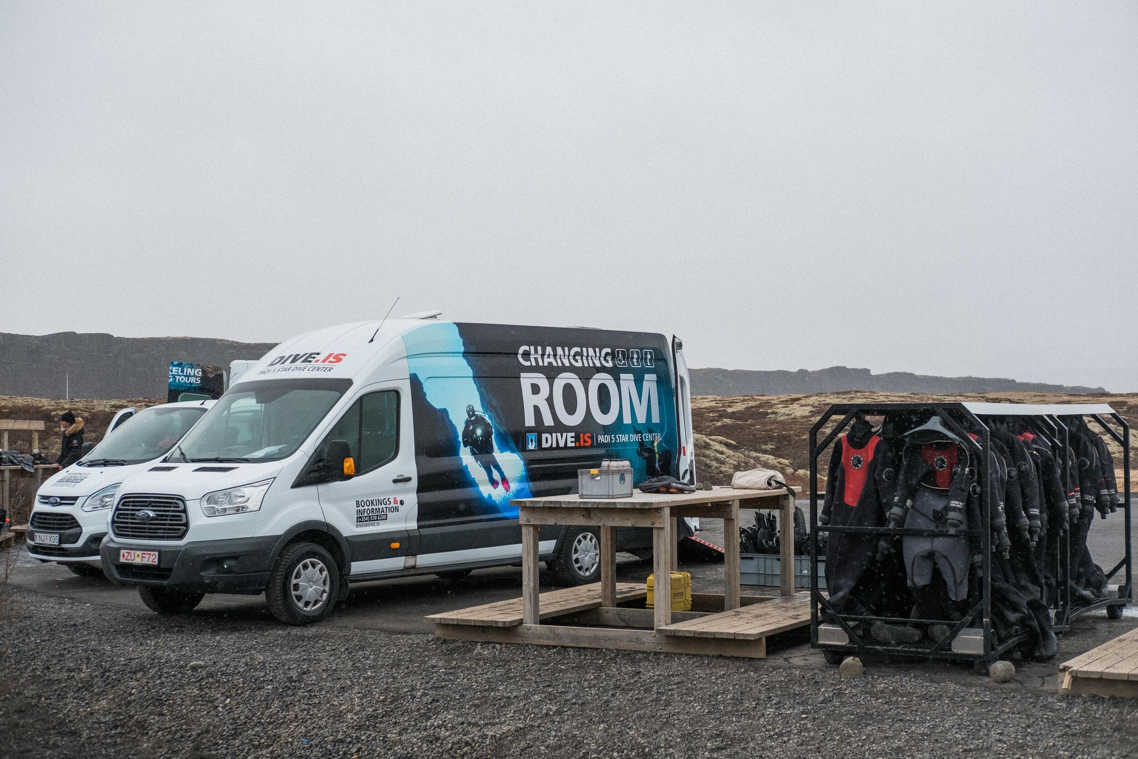 A white DIVE.IS van with "CHANGING ROOM" branding, parked next to a rack of drysuits in an outdoor, rocky landscape.