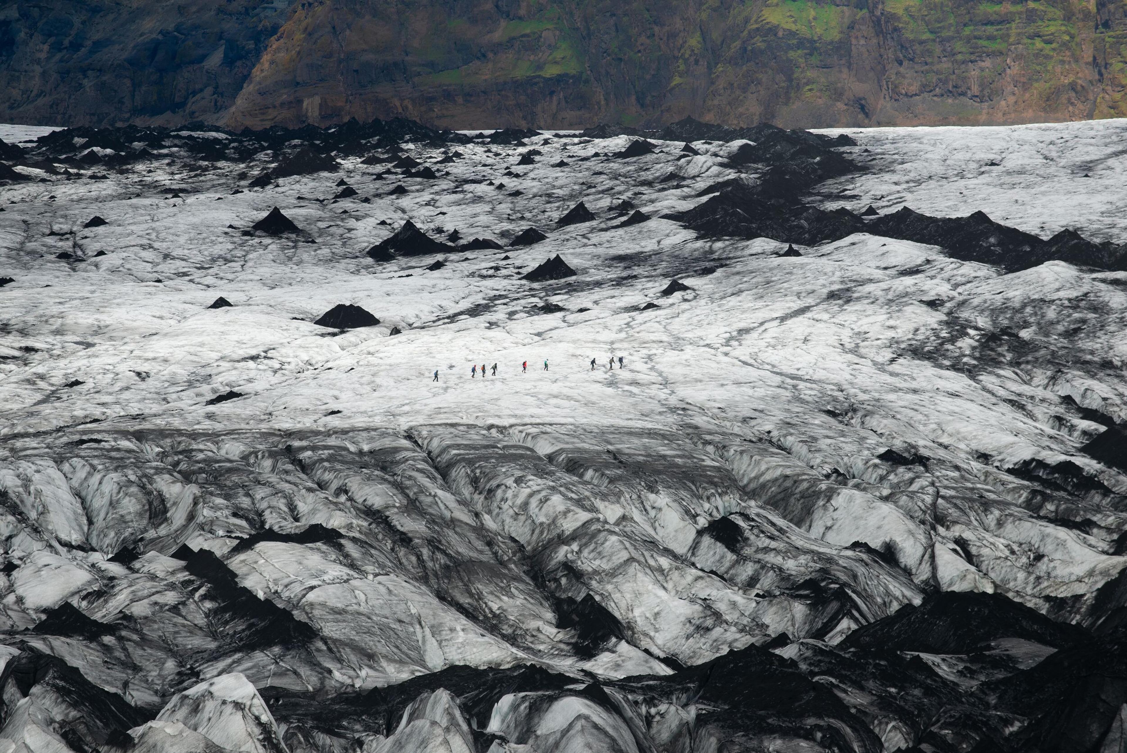 A small group of hikers traverses the vast, rugged landscape of Sólheimajökull glacier, surrounded by black volcanic ash and ice. A dramatic Sólheimajökull glacier hike showcasing Iceland’s unique terrain.