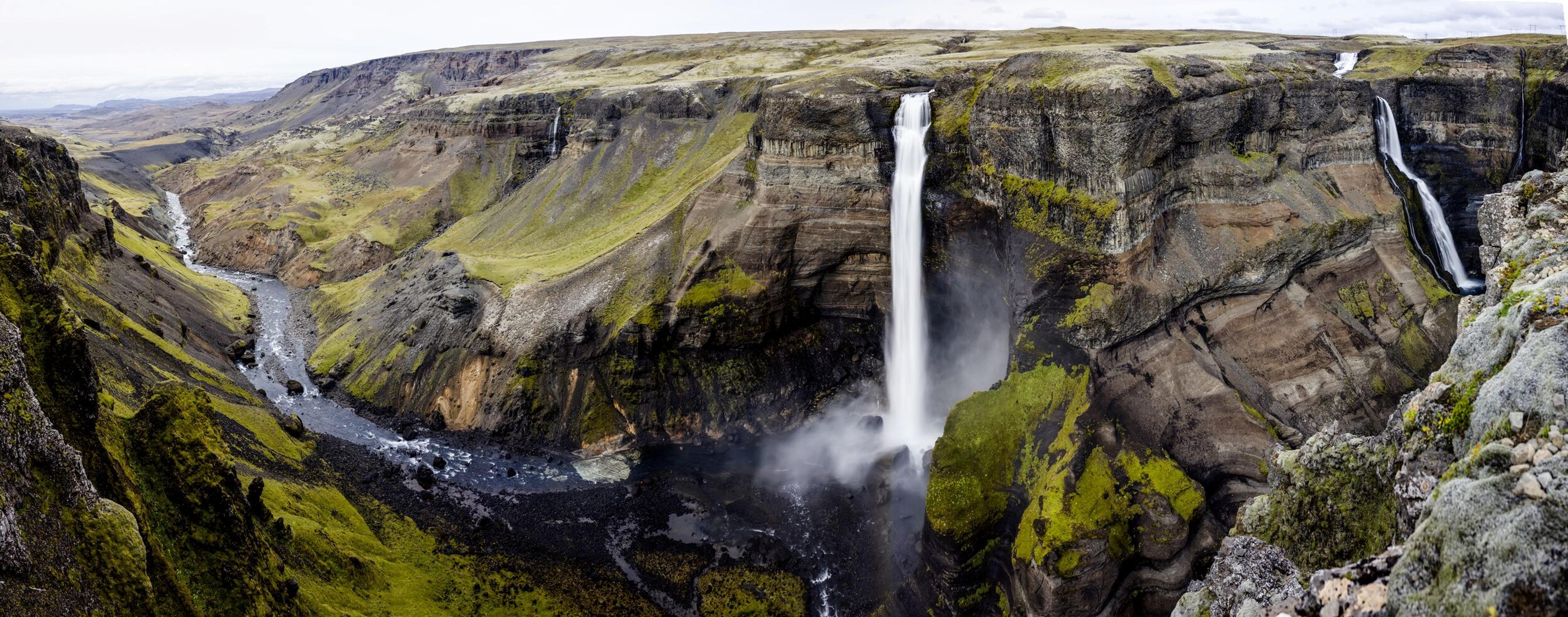Panoramic view of a deep, mossy canyon with a river and two waterfalls, one very tall.