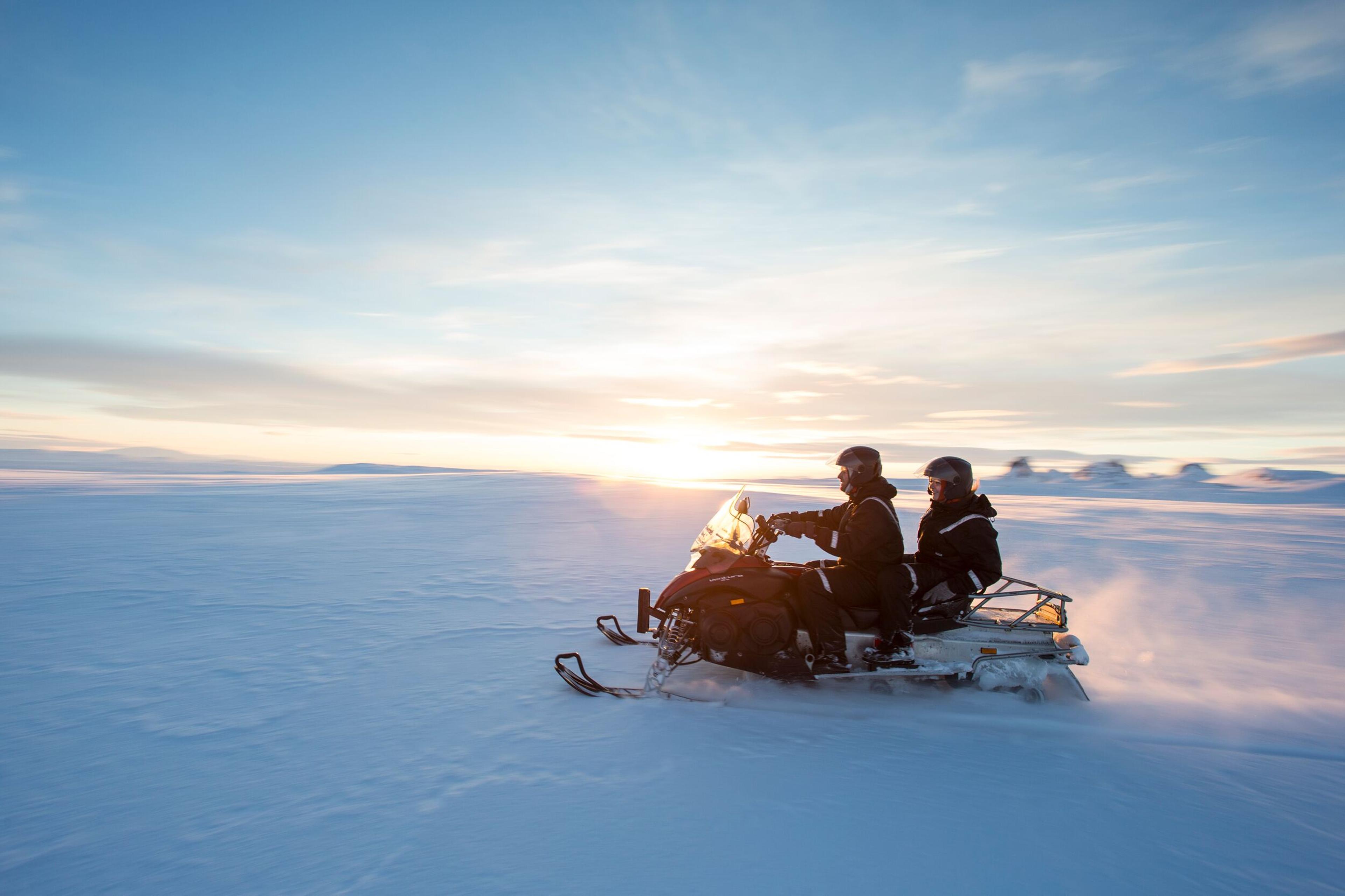 Two individuals riding a snowmobile across a vast glacier, silhouetted against the radiant glow of a setting sun.