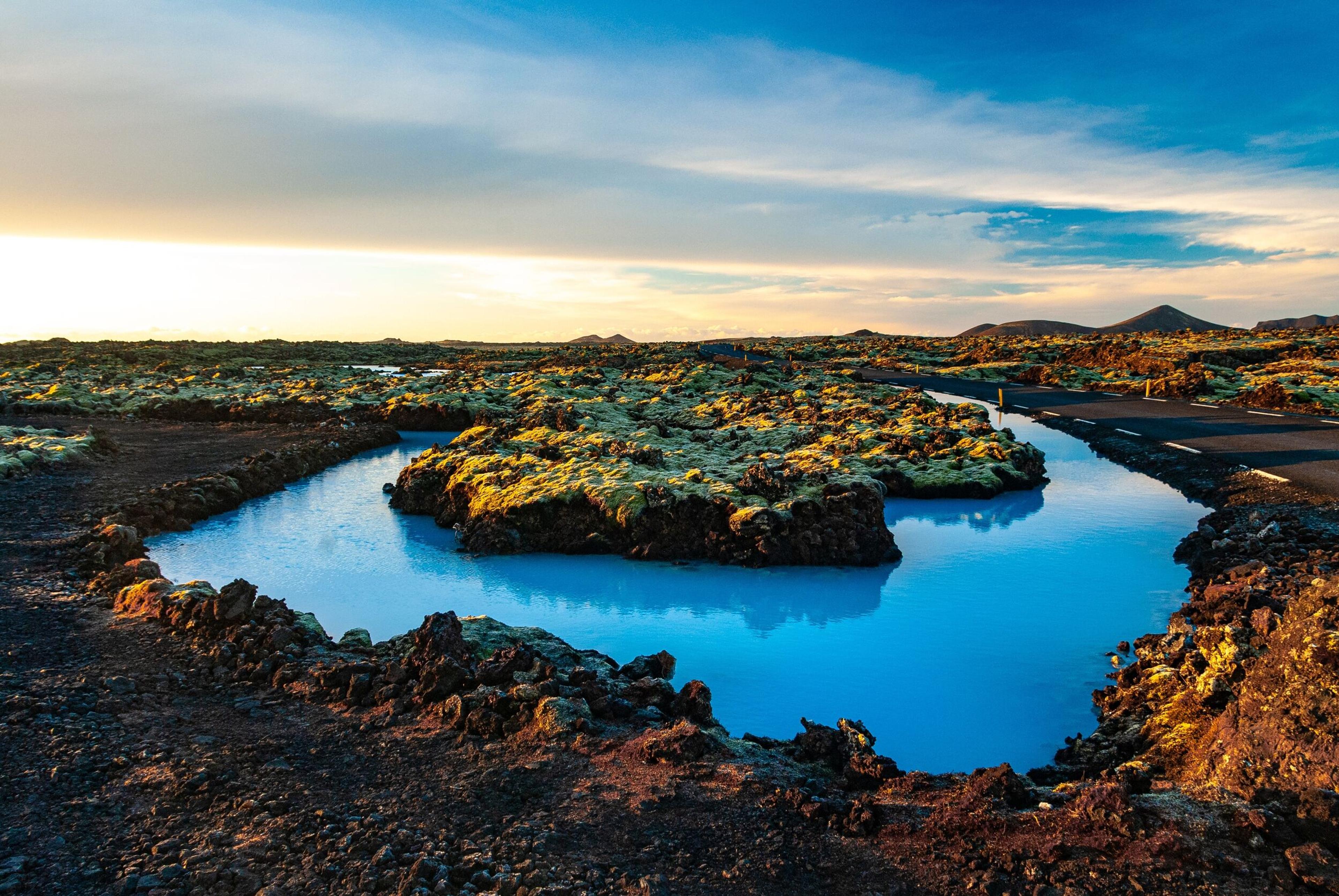 Vibrant blue geothermal water winding through mossy lava fields with a road under a bright sky.