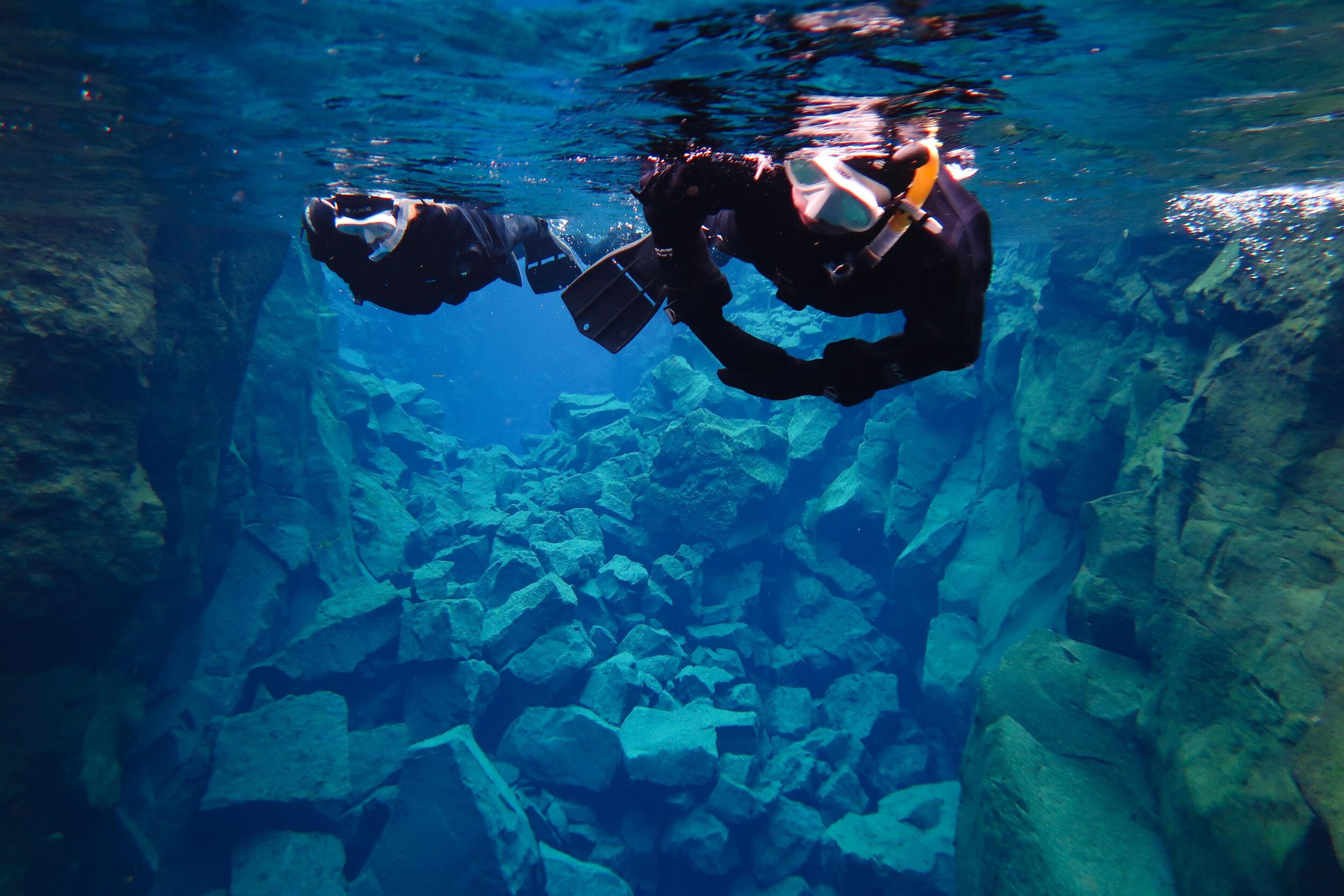 A snorkeler in a wetsuit floats in incredibly clear blue water above a rocky underwater canyon.