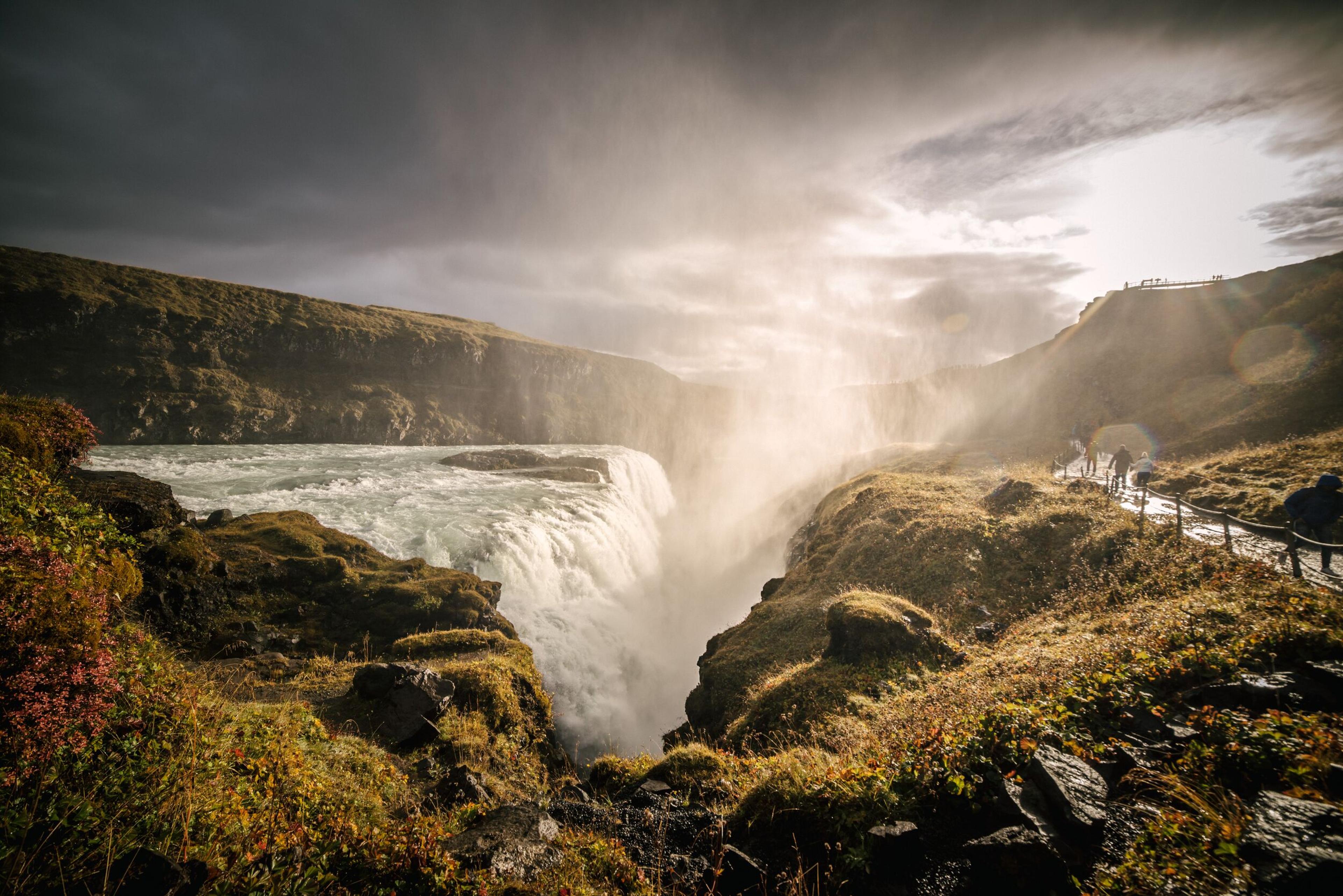  A breathtaking view of Gullfoss, a famous waterfall in Iceland. The powerful, cascading water flows in two tiers down a rugged canyon, surrounded by lush greenery. Mist rises from the falls, creating a soft, ethereal haze.