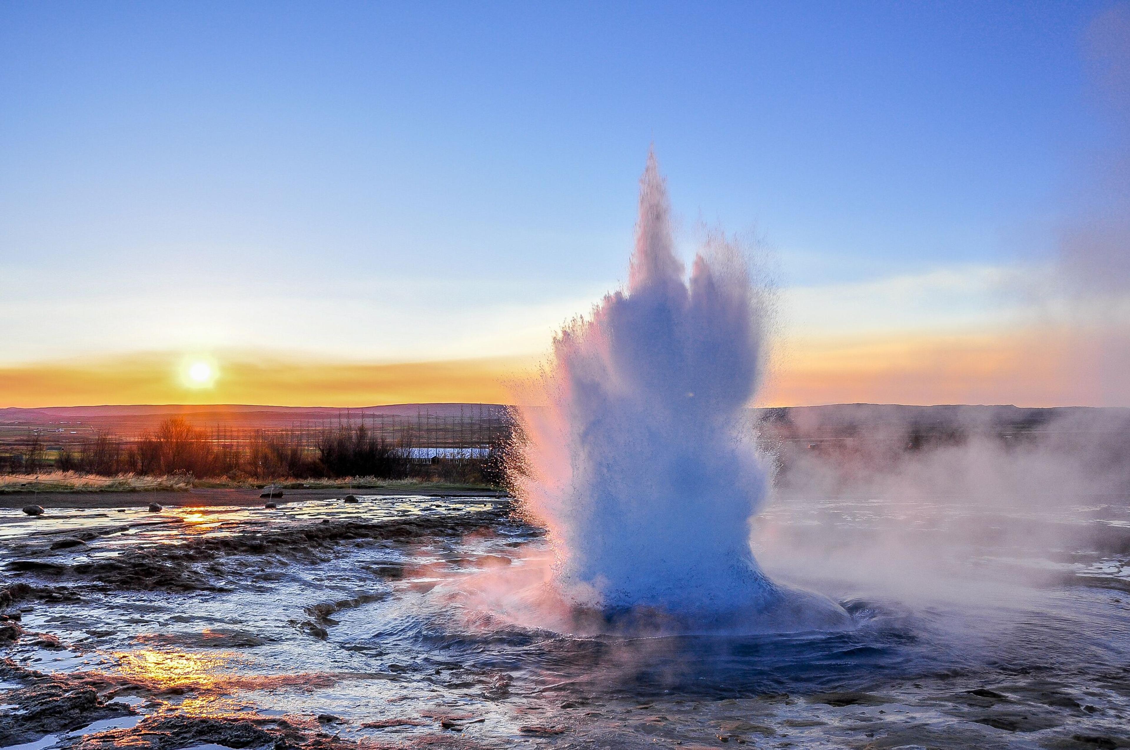 : A vibrant geyser erupts with water and steam at sunrise, set against a clear blue sky with a warm glow on the horizon. Surrounding terrain reflects the light, emphasizing the unique geothermal activity.